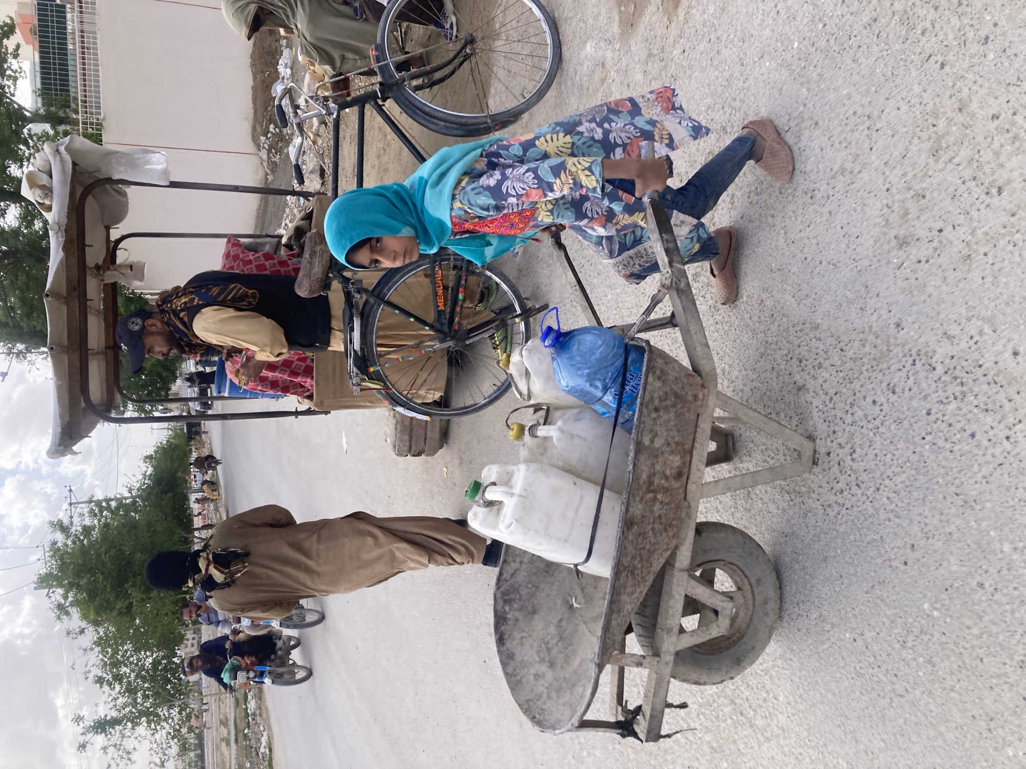 In Quetta City Tehsil, Pakistan, a young girl wearing a blue headscarf and floral patterned dress stands next to a cart filled with water containers. The cart is connected to a bicycle, beside which a man is standing. A second man is seen pushing the cart, all on a paved road. In the background, there are buildings, trees, and other people on motorcycles. The environment appears to be a residential area.