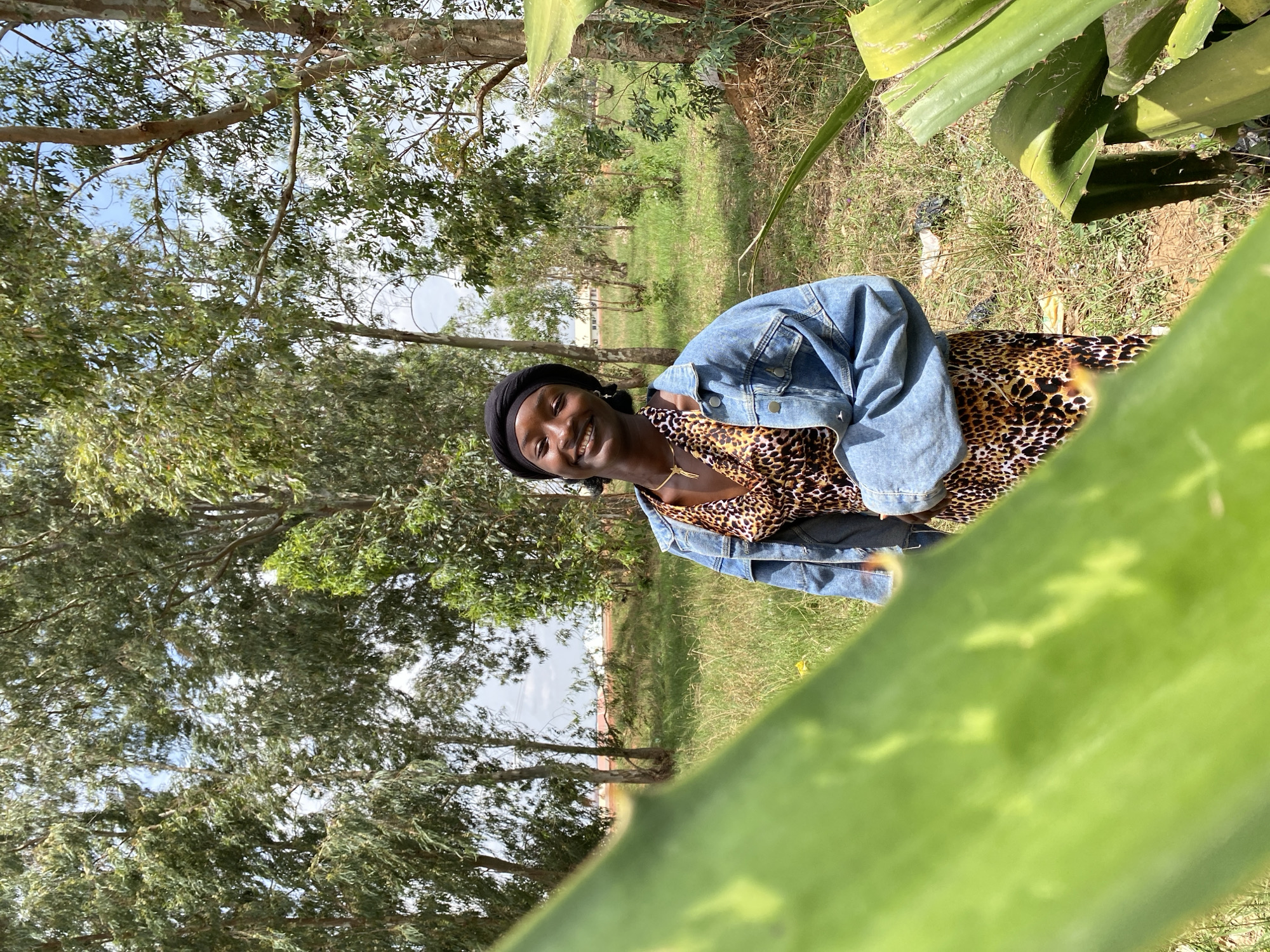 The image shows a woman standing outside in a grassy area with trees. She is wearing a black head covering, a leopard-print dress, and a blue denim jacket. She is smiling and looking at the camera. The background includes trees, grass, and a slightly visible horizon line. The location is likely in a rural or semi-rural area. There is no visible evidence in the image to suggest that the location is in the city of Durimi, Nigeria. There is no city visible in this image.