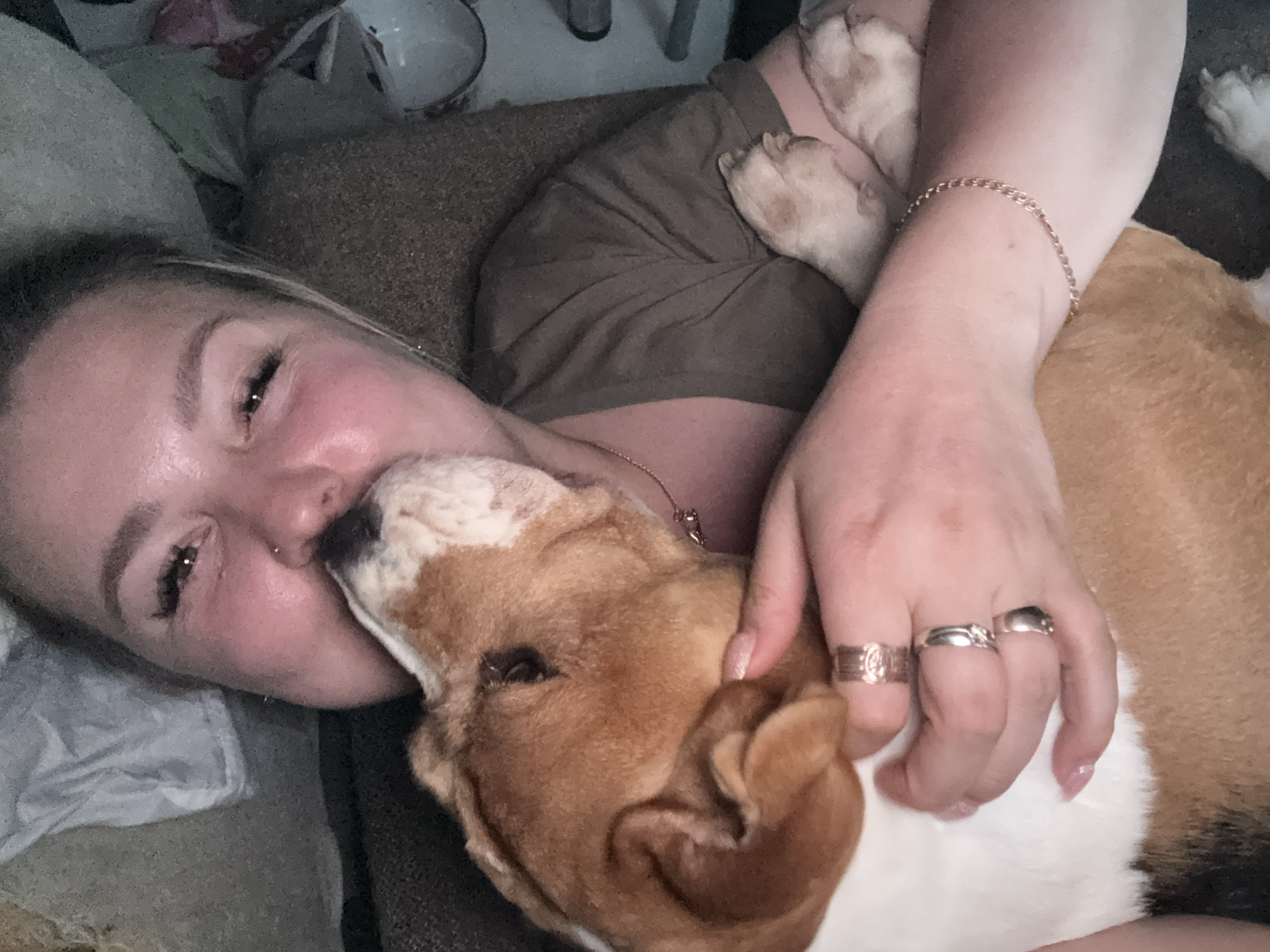 A woman is lying down, embracing a tri-colored beagle dog. The dog is partially in front of her and she is kissing its nose. The woman is fair-skinned with blonde hair and is wearing multiple rings on her right hand and a chain bracelet on her right wrist. She is wearing a dark-colored t-shirt. The dog has its white paws near her right shoulder. The setting appears to be indoors on a dark-colored cushioned surface. The location is not discernible from the image, other than it might have taken place in Russia.