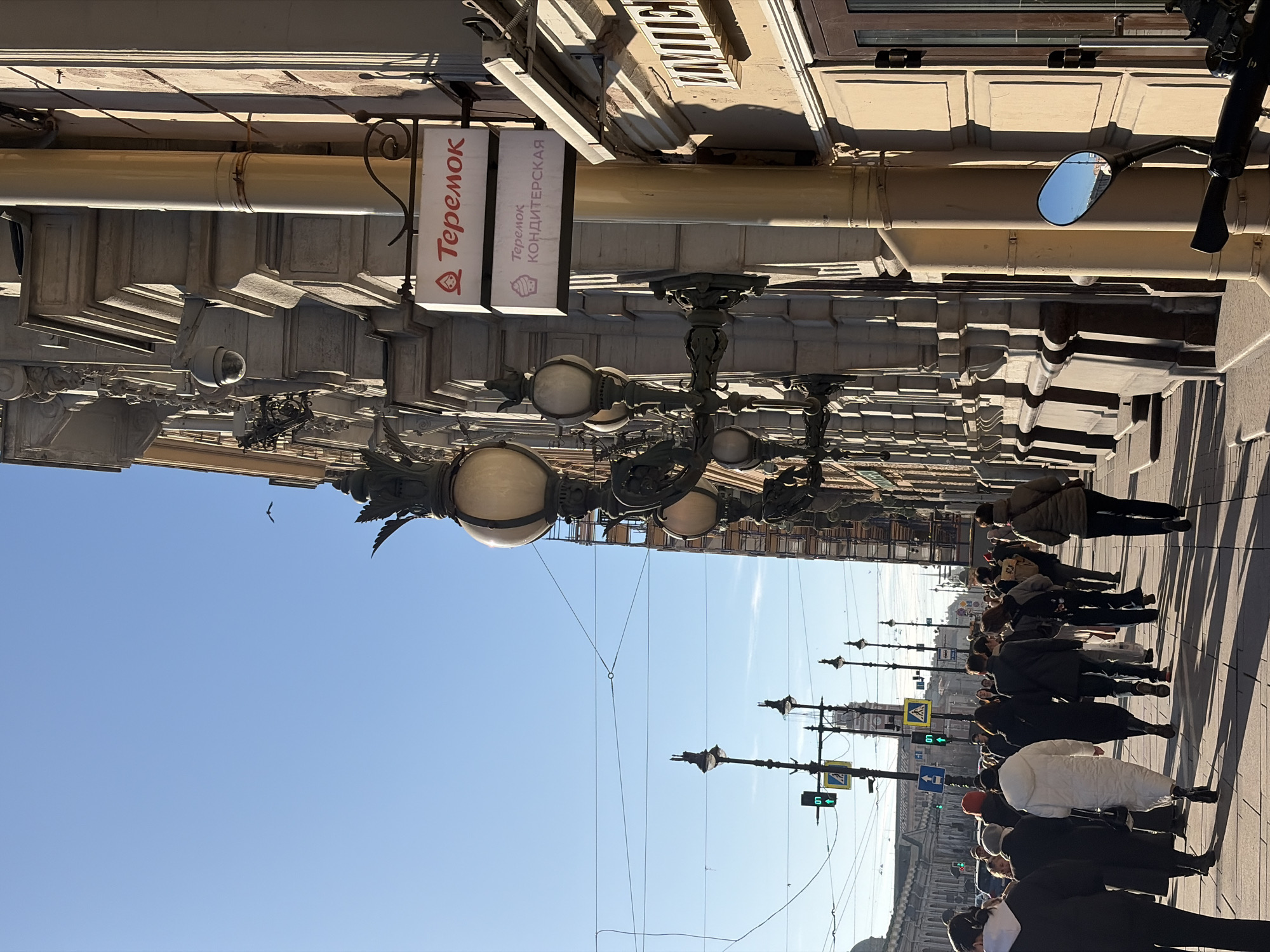 The image shows a street scene in Saint Petersburg, Russia, viewed from a slightly high perspective and rotated 90 degrees counter-clockwise. A group of people is walking along a sidewalk. Above the sidewalk are decorative street lamps with spherical globes. A sign for "Teremok Konditerskaya" (Teremok Confectionery) is visible. The sky is clear and blue. A small bird is flying in the sky near the building. A motorcycle mirror is visible on the building on the right. Traffic lights are visible and display green lights. The image is taken from an elevated position, likely from a building or higher vantage point. The building has elaborate architectural details.
