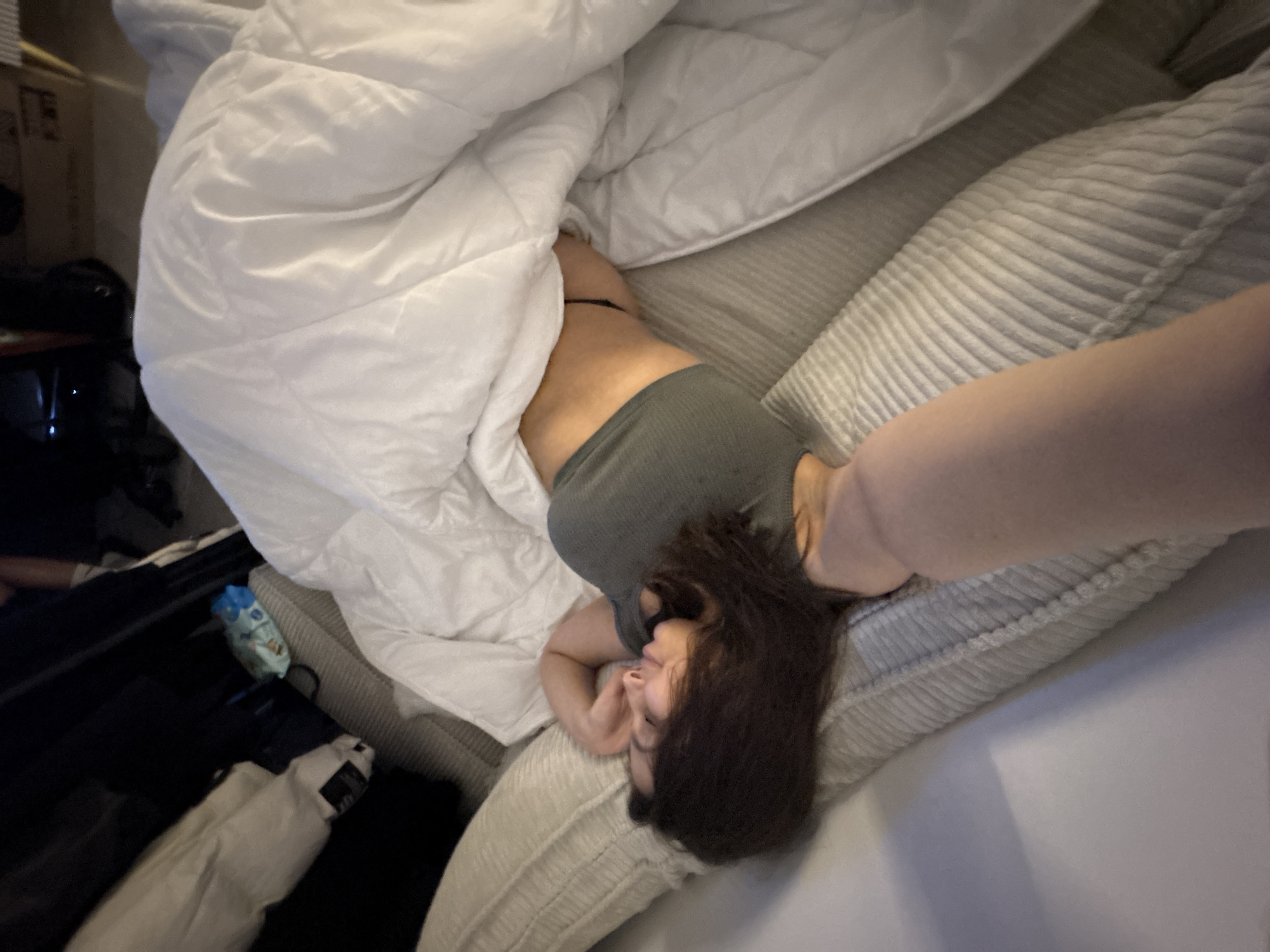 A woman lies on a striped light gray sofa partially covered by a white duvet. She wears a gray top and is partially under the covers. Her dark hair is spread on the sofa cushion. Beside the sofa is a black suitcase with a white bag on top. A cardboard box is visible in the background. The location cannot be determined from the image.