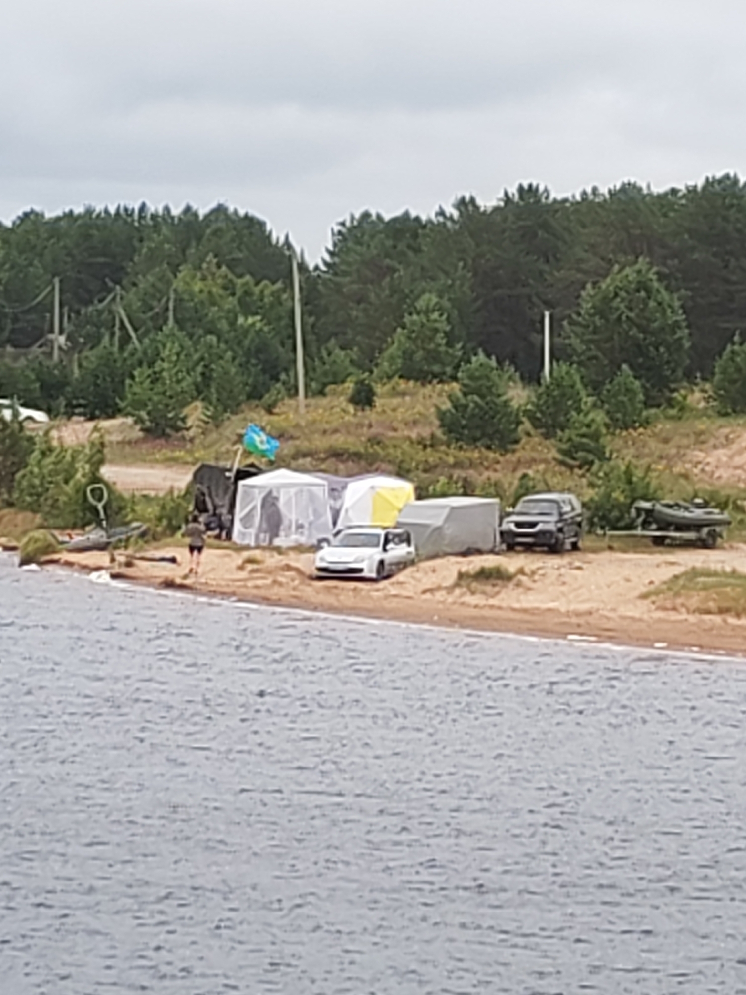 In Shalsky, Russia, on a sandy shore adjacent to a body of water, a campsite is established. Several tents, one white and another with yellow accents, are erected on the sand. A dark-colored SUV is parked behind the tents, and a white car is parked to the left of the tents. A military-style vehicle is positioned behind a blue and green flag. To the right of the SUV, a boat is on a trailer. A single person, wearing a dark shirt and shorts, is walking away from the water towards the campsite. The background features a dense line of coniferous trees. The sky is overcast.