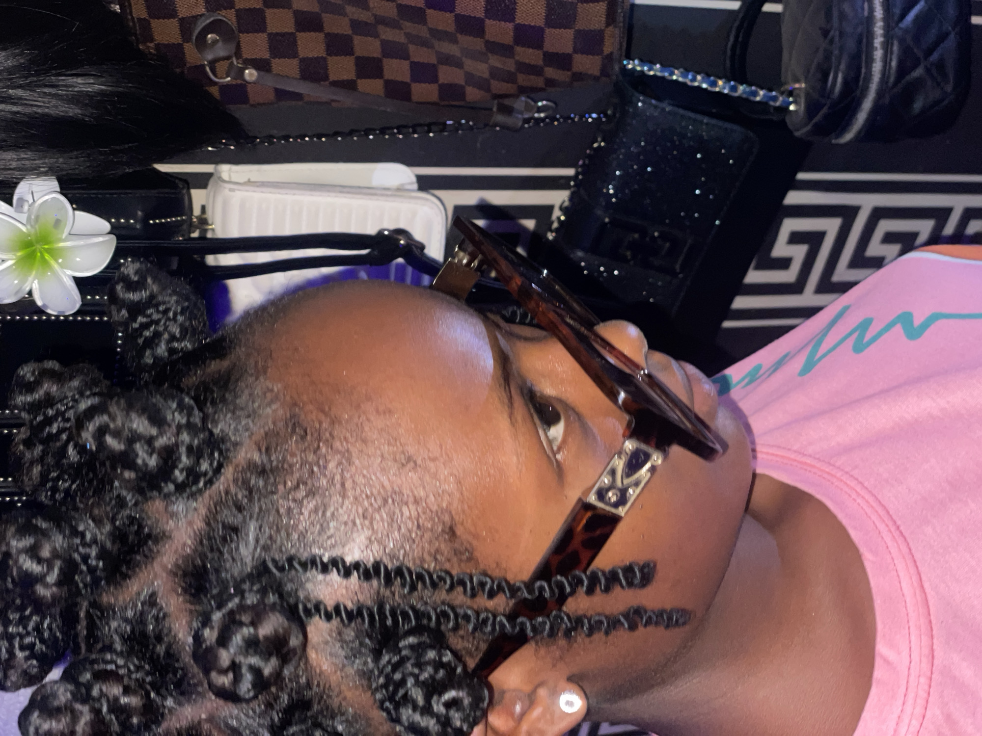 The image depicts a Black person with dark hair styled in coiled knots, wearing tortoiseshell glasses. They are positioned against a backdrop of multiple purses and a striped surface. The subject wears a pink shirt. There are no indications this photo was taken in Lokoja, Nigeria.