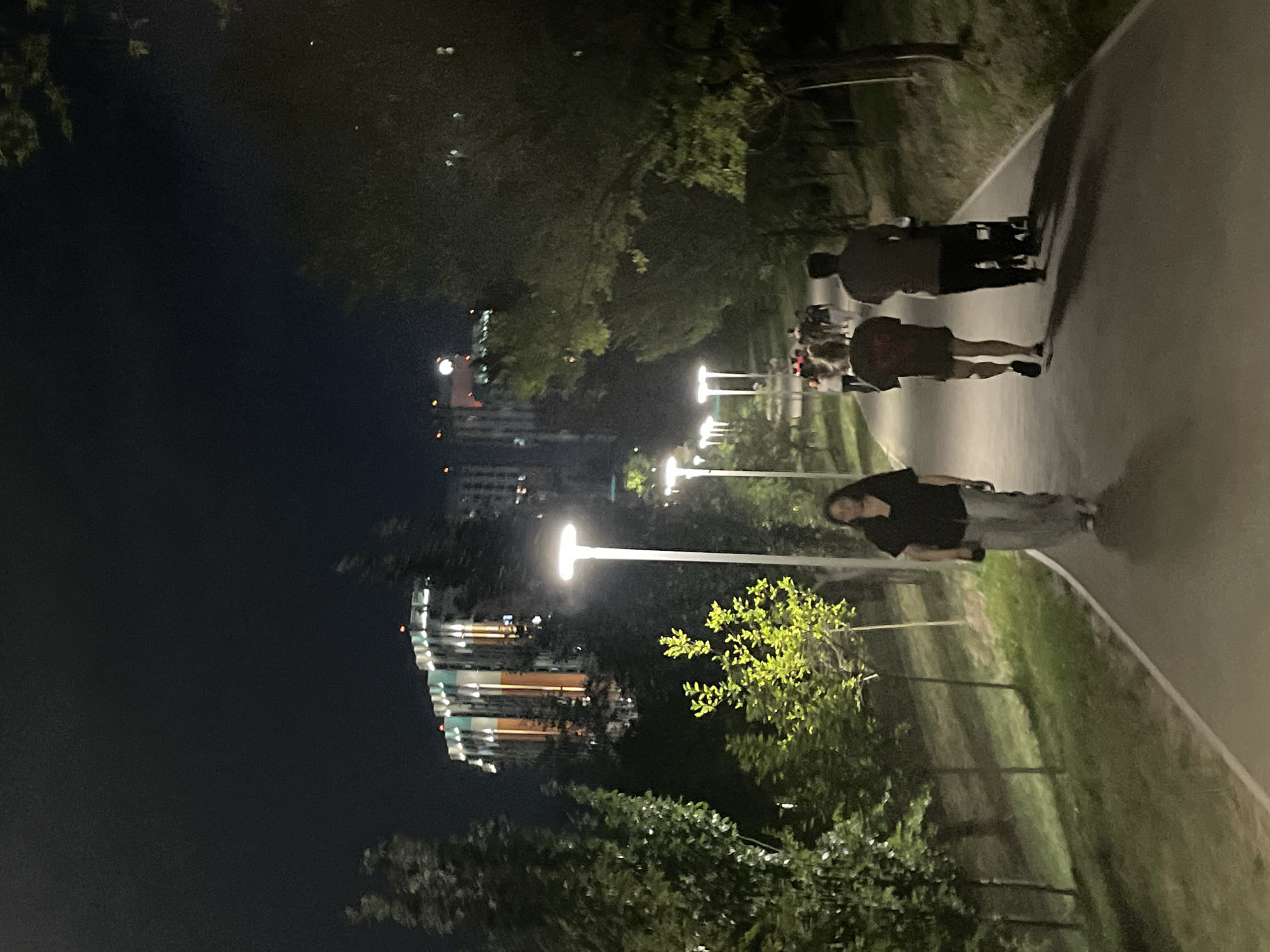 This is a nighttime photograph of a park pathway in Almaty, Kazakhstan. A group of people are walking along the path, illuminated by streetlights. In the background, a modern building with illuminated windows is visible. The pathway is paved and bordered by trees and grassy areas. The sky is dark, indicating nighttime.