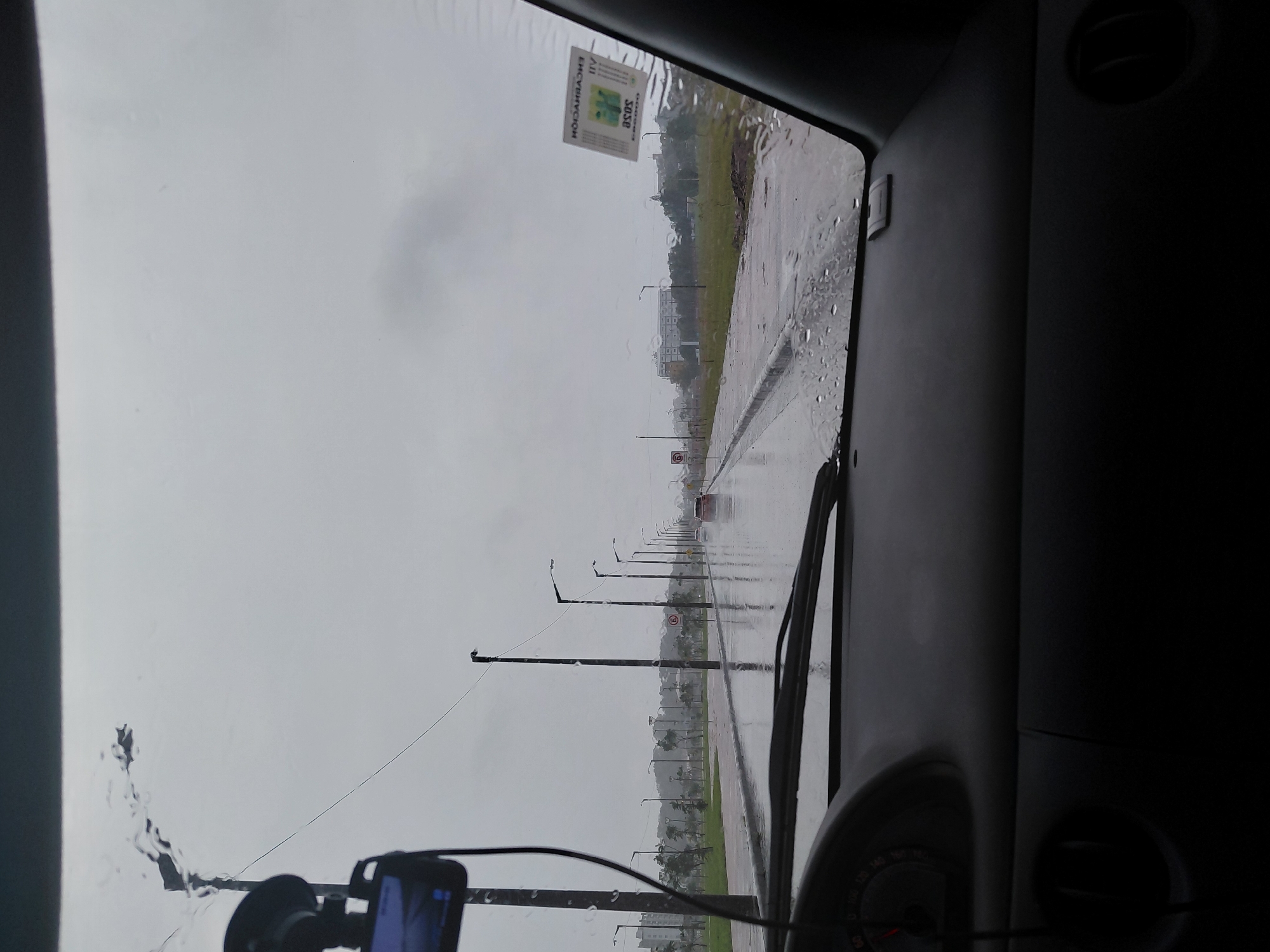 The image depicts a view from inside a vehicle looking out through a rain-streaked windshield onto a wet, urban street. The sky is overcast and grey, indicating rainy weather. A row of streetlights lines the right side of the road, with reflections visible on the wet asphalt. Several vehicles are present on the road, appearing as distant blurs.
A sticker is affixed to the windshield on the passenger side. The sticker is rectangular and has text and graphics on it. The visible text on the sticker includes "ENCARNACION", "III", and "asos".
The interior of the vehicle is visible in the foreground, showing the dashboard, part of the steering wheel, and a vent. The overall scene suggests a drive through the city of Encarnacion, Paraguay, during a downpour.