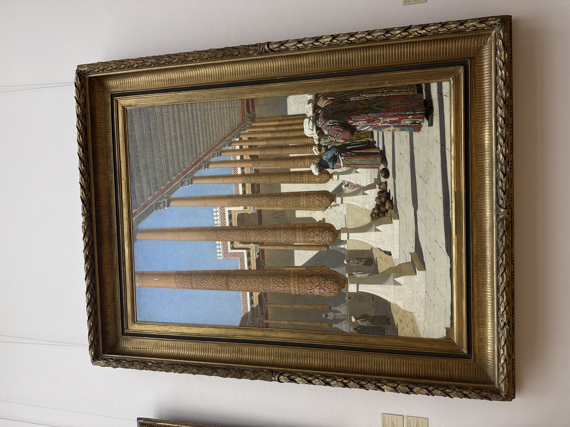 The image depicts an artwork in a gilded frame, hanging on a white wall. The artwork appears to be a painting or illustration of an ornate, open-air structure featuring numerous tall, decorative columns. People are present within the scene, some wearing traditional clothing or turbans. The architecture suggests a historical or exotic setting. There is also a shadow in the bottom right corner. The artwork is located in Moscow, Russia.
