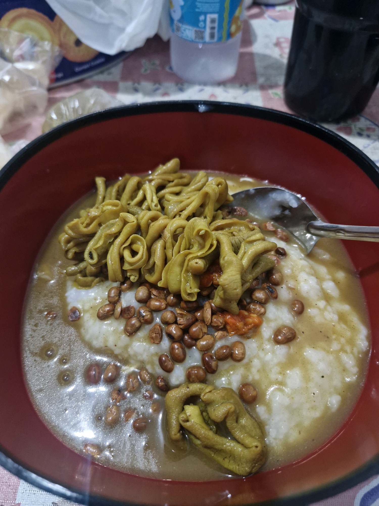 A close-up overhead shot shows a bowl of bubur ayam (chicken porridge) in Rantau, Indonesia. The porridge is served in a red bowl with a black rim, and it is topped with a generous portion of fried intestines, peanuts, and chili paste. A spoon rests in the bowl, partially submerged in the porridge. The background is out of focus, but it appears to be a table with a patterned tablecloth, a plastic bag, and a dark beverage container. The lighting is dim, suggesting the photo was taken indoors, possibly in a restaurant or home.
