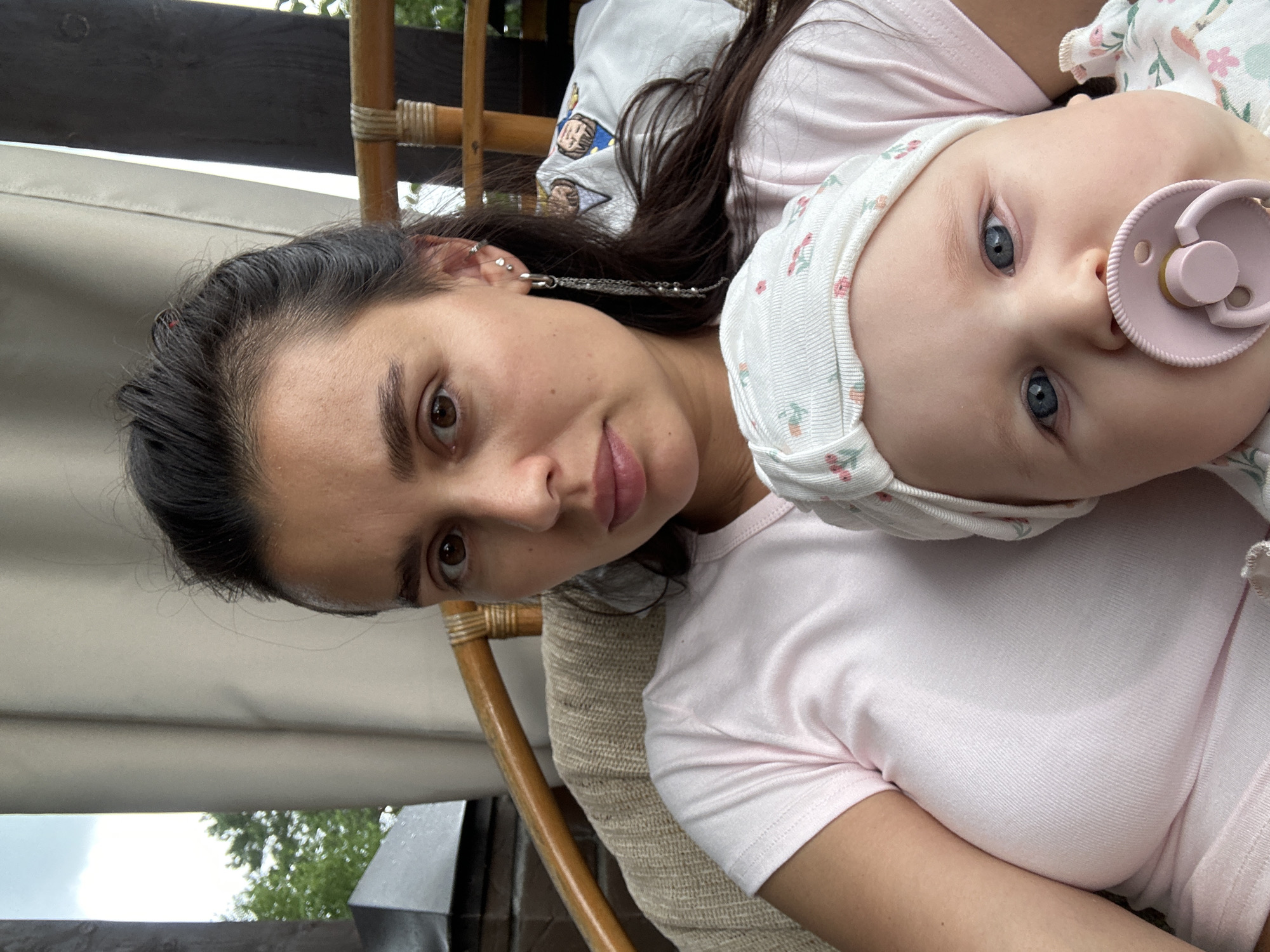A mother and baby are posing for a selfie outdoors, likely on a covered porch or balcony in Sylva, Russia. The mother, with dark hair and brown eyes, is wearing a light pink t-shirt and a dangling earring. She holds the baby, who has blue eyes and is wearing a white hat with a floral pattern and a pink pacifier. The background shows a wooden railing, beige curtains, and greenery, suggesting an outdoor setting. The lighting is natural and appears to be daytime.