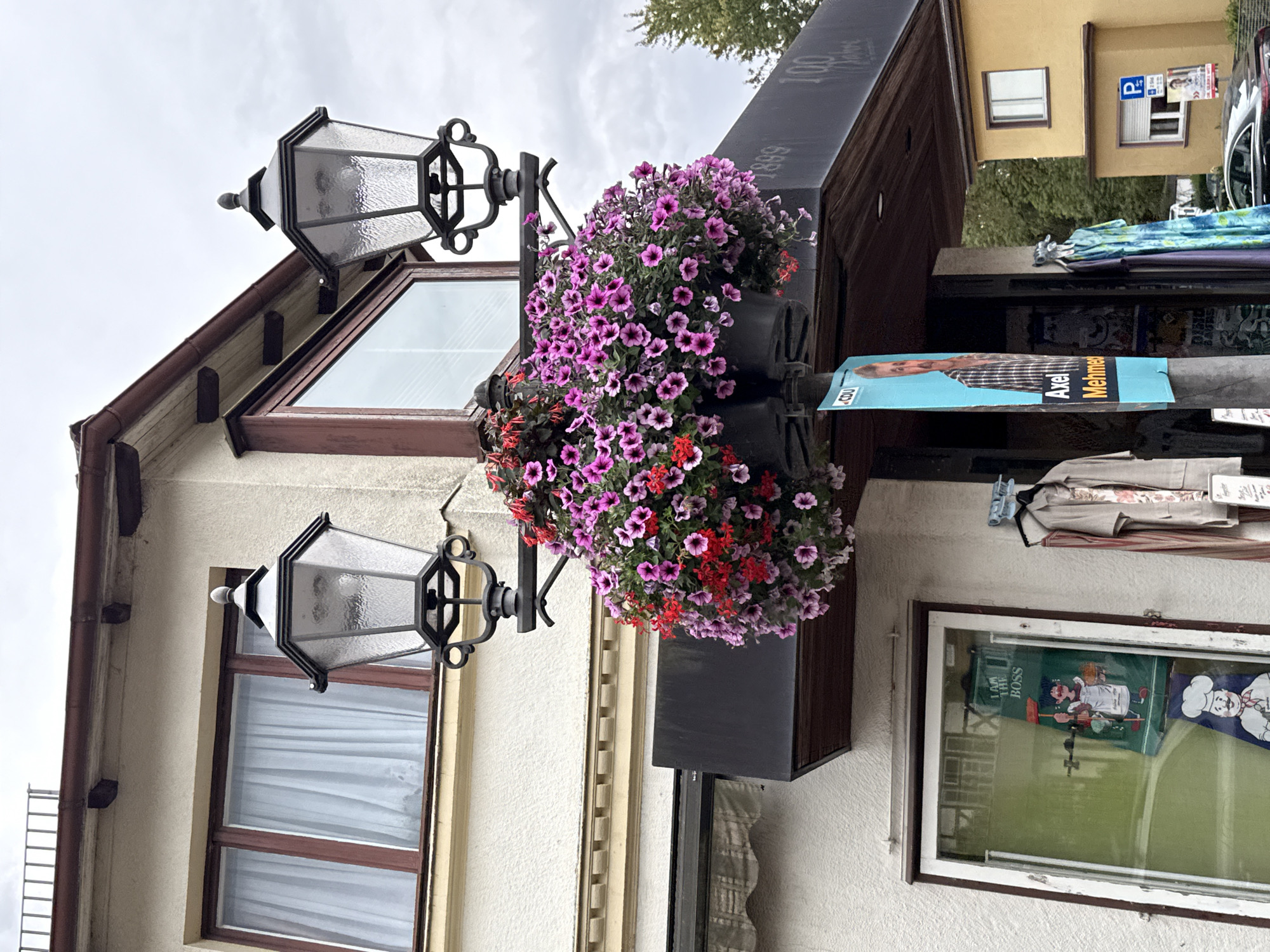 This is a close-up, eye-level, outdoor, daytime shot of a storefront in Bad Sachsa, Germany. The storefront is a light-colored building with a dark awning and a large window displaying an advertisement for a baker and a chef. Above the awning, a lamppost is adorned with hanging baskets of pink and red flowers. Two antique-style lanterns are attached to the lamppost. To the right of the storefront, a yellow building is visible, and a parked car is in the foreground. A blue poster with a man's face and the name "Axel Mehme" is attached to the lamppost.