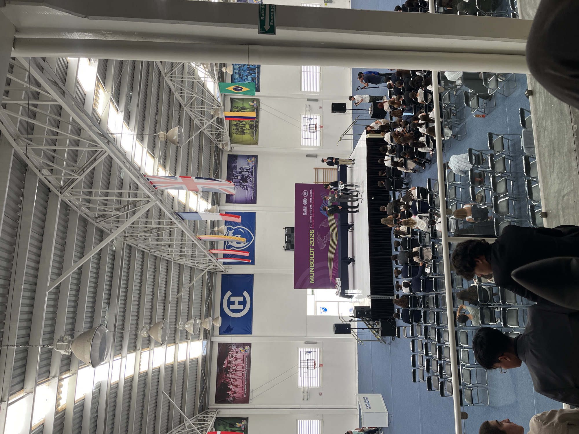 The image depicts an indoor event, likely an assembly or conference, taking place in a large gymnasium or multi-purpose hall. The perspective is from an elevated vantage point, looking down and across the space.
The scene features a stage at the far end of the hall, where several individuals are gathered, one standing at a podium addressing an audience. The audience is seated in numerous rows of chairs on a blue floor, facing the stage. Many seats in the foreground are empty, while the bulk of the attendees are visible closer to the stage.
The hall has a high, white metal-trussed ceiling with several skylights providing natural daylight, suggesting the event occurs during daytime. Large industrial pendant lights are also suspended from the ceiling. Decorations include multiple national flags hanging vertically from the ceiling structure, notably the Union Jack (United Kingdom), the flag of Brazil, and the flag of Colombia. Various banners adorn the walls, including one with the text "Un equipo, una familia, una pasión" (A team, a family, a passion) and another large blue banner featuring a stylized white "H" logo.
The main backdrop banner on the stage reads "MUNBOLDT 2026 Building bridges, not walls," strongly suggesting a Model United Nations event. A white sign or box in front of the stage displays "COLEGIO HUMBOLDT." On an upper wall, a green "Ruta de Evacuación" (Evacuation Route) sign with an arrow is visible. Basketball hoops on the walls further confirm the gymnasium setting.
The event is held in Puebla, Mexico, as indicated by the provided location context.