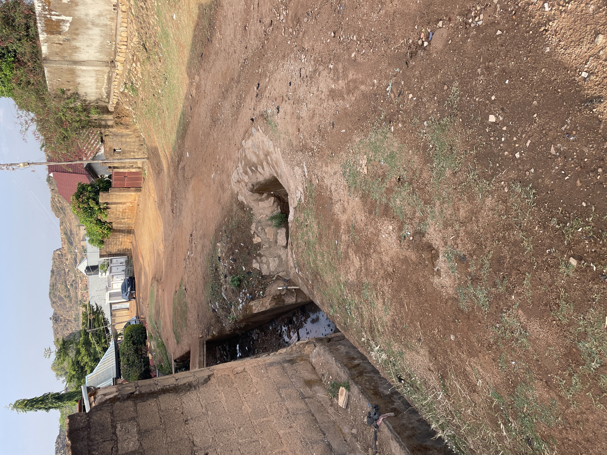 The scene depicts an unpaved street in Jos, Nigeria, characterized by a dirt path and a drainage channel. A partially visible concrete structure surrounds a hole leading into the drainage system. The channel contains stagnant water with visible debris and trash. Adjacent to the street, there are residential buildings, including structures with corrugated metal roofing and brick walls. The environment includes sparse vegetation growing in the dirt and buildings in the distance that are situated on a mountain.