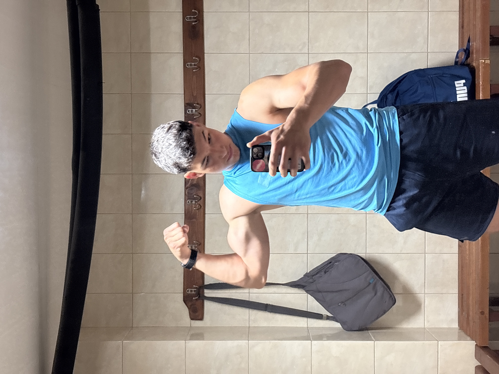 A young man with short, light-colored hair stands in a locker room and takes a mirror selfie with his phone. He is wearing a bright blue sleeveless shirt and dark blue shorts. His left arm is flexed, showing his bicep. A grey duffel bag is hanging on a hook to his left, and a blue backpack with a white logo is on the bench behind him. The walls are tiled in a beige color, and a wooden bench runs along the bottom of the frame. The scene appears to be within the Zlatograd Sports Hall locker room.