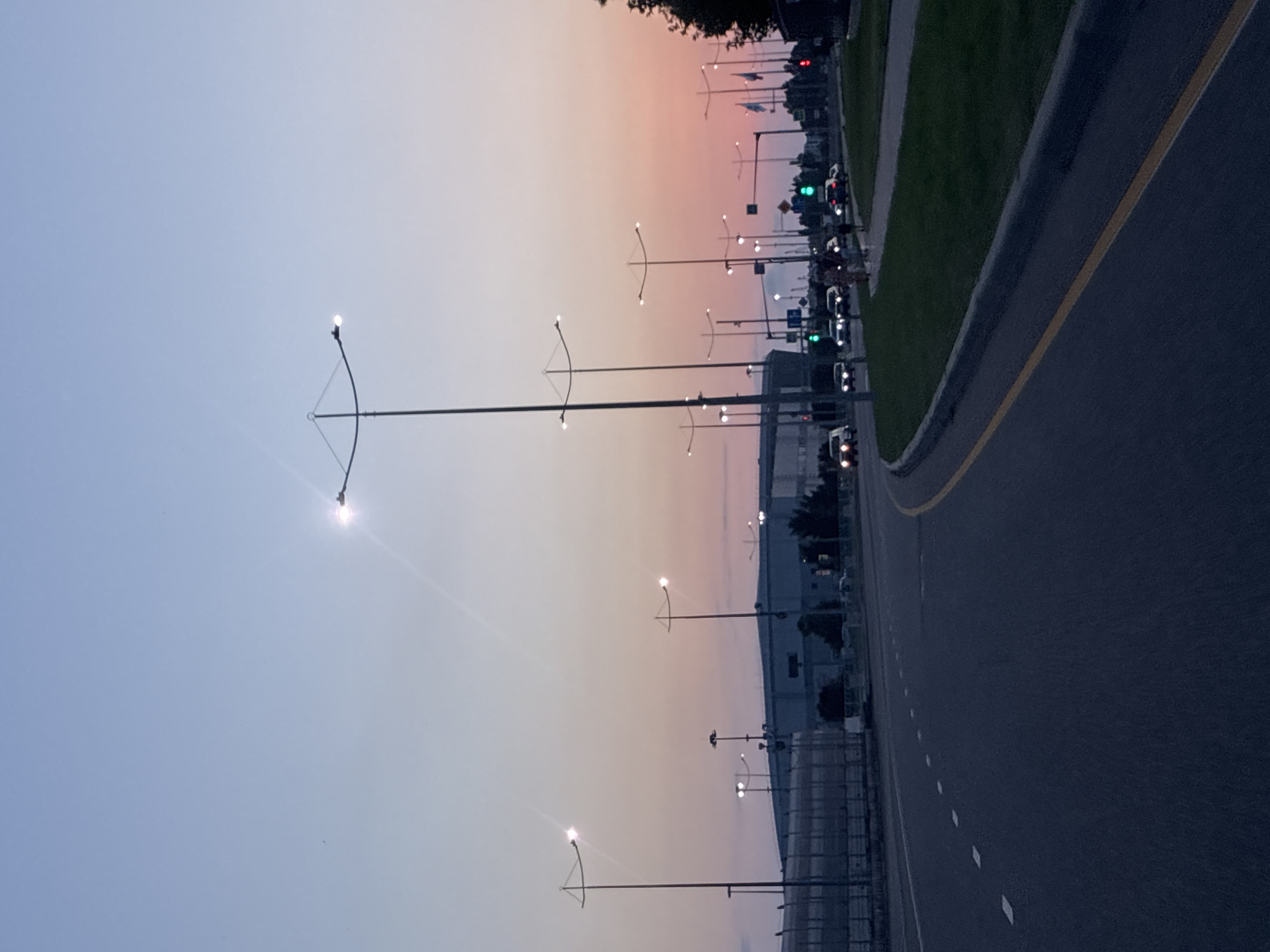 The image shows a street scene in Sirius, Russia, during twilight. The sky transitions from a pale blue at the top to a warm pink and orange hue near the horizon, indicative of sunset. A multi-lane road curves into the distance on the left side of the frame, marked with white dashed lines. A yellow line separates the lanes of traffic. On the right side of the road, there is a grassy area and a sidewalk.
Several streetlights with distinctive designs, featuring curved arms holding lights, are visible along the road. Some of these lights are illuminated. In the background, there is a large, modern-looking building, possibly an arena or exhibition center, with a long, flat roof. Further in the distance, other structures and trees can be seen.
The road is moderately busy with traffic. Several cars are visible, some with their headlights on. A traffic light in the distance shows a green signal. A few pedestrians are walking on the sidewalk on the right side of the road. The overall impression is one of an urban or suburban environment at the end of the day.