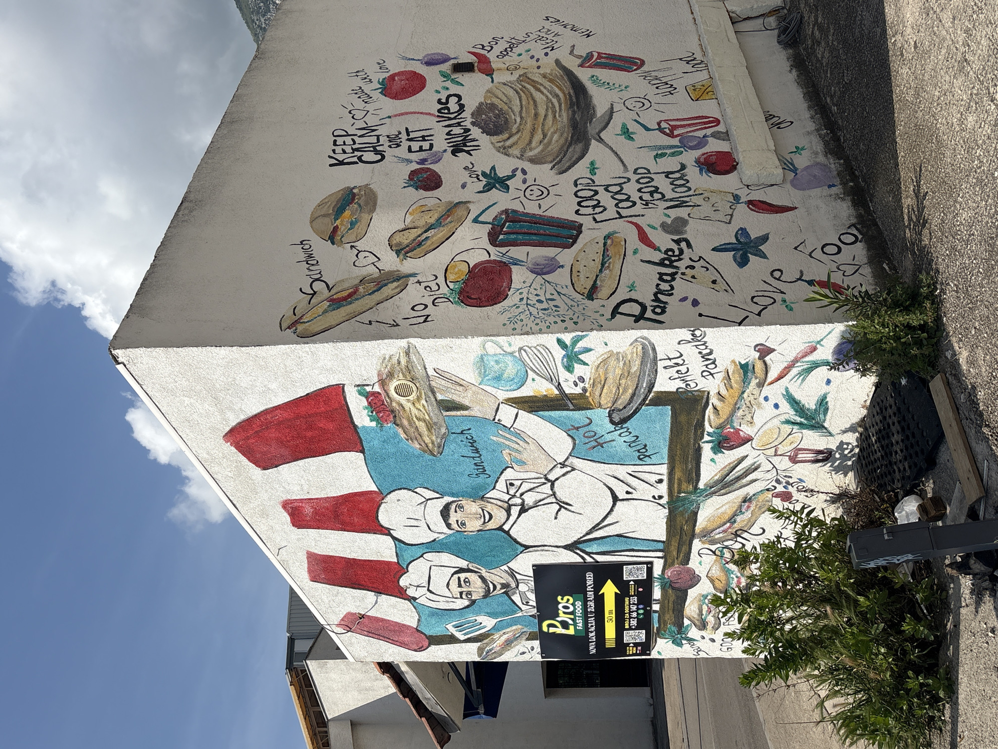 An outdoor scene captures a building wall adorned with vibrant murals, presented as if the camera was tilted 90 degrees counter-clockwise. The bright daylight illuminates a blue sky with fluffy white clouds visible in the upper left.
The wall is divided into two main mural sections. The upper section is a colorful collage of various food items such as pancakes, sandwiches, tomatoes, onions, and chili peppers, alongside kitchen utensils and positive affirmations. Visible text includes "KEEP CALM and EAT PANCAKES," "Sandwich," "No Diet," "Bon appetit," "Meals And Memories," "Good Food Good Mood," "Happy," and "LOVE." A large, stylized spiral pancake or pastry is a prominent element.
The lower mural features two cheerful cartoon-style chefs in white uniforms and hats, actively preparing food. One chef holds a spatula, and the other presents a dish. They are set against a blue and red background, surrounded by more food illustrations like baguettes, strawberries, and other ingredients. Text here includes "Sandwich," "Hot Pancake," and "Perfekt Pancakes."
In the foreground, a black sign with yellow text for "Pros FAST FOOD" is visible, partially obscured by green bushes. The sign directs "NOVA LOKACIJA U ZGRADI PORED" (New Location in the Building Next Door) "50 m" with an arrow pointing right. It also provides a "BROJ ZA DOSTAVU" (Delivery Number): "+382 66 147 233." The ground next to the wall is concrete, with some weeds and a drain cover. The overall scene suggests a lively, food-themed establishment.