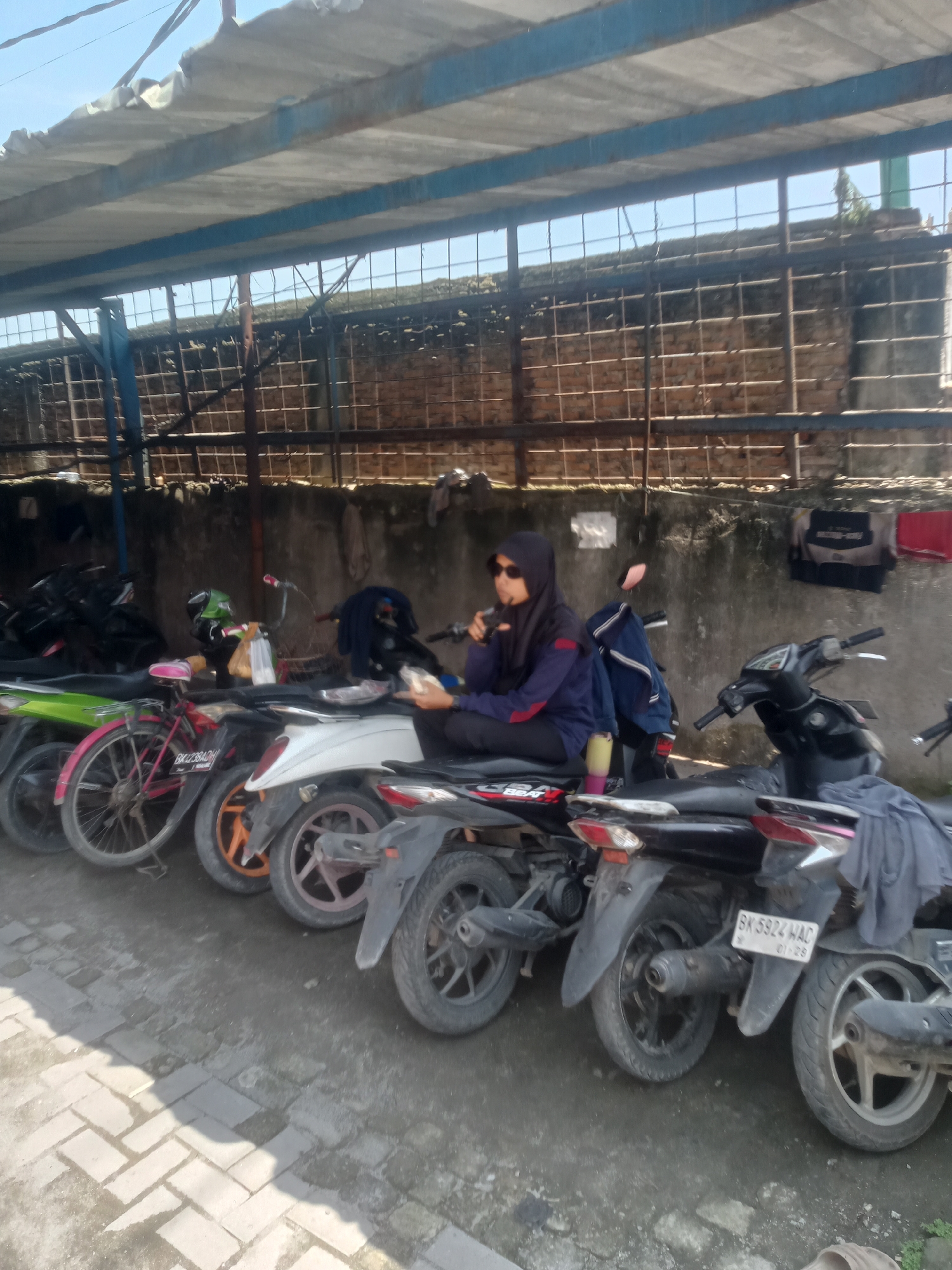 In Sunggal, Indonesia, a person wearing a black hijab and sunglasses is seated on a black motorcycle under an open-air parking structure. The person is holding a cup and appears to be drinking. A blue backpack is behind them on the motorcycle. A variety of other motorcycles are parked in a row next to them, including a pink bicycle. The background consists of a concrete wall and a fenced enclosure. The ground is paved with interlocking bricks. Laundry items are hanging on the wall in the background. The scene suggests a parking area in a residential or commercial district of Sunggal.