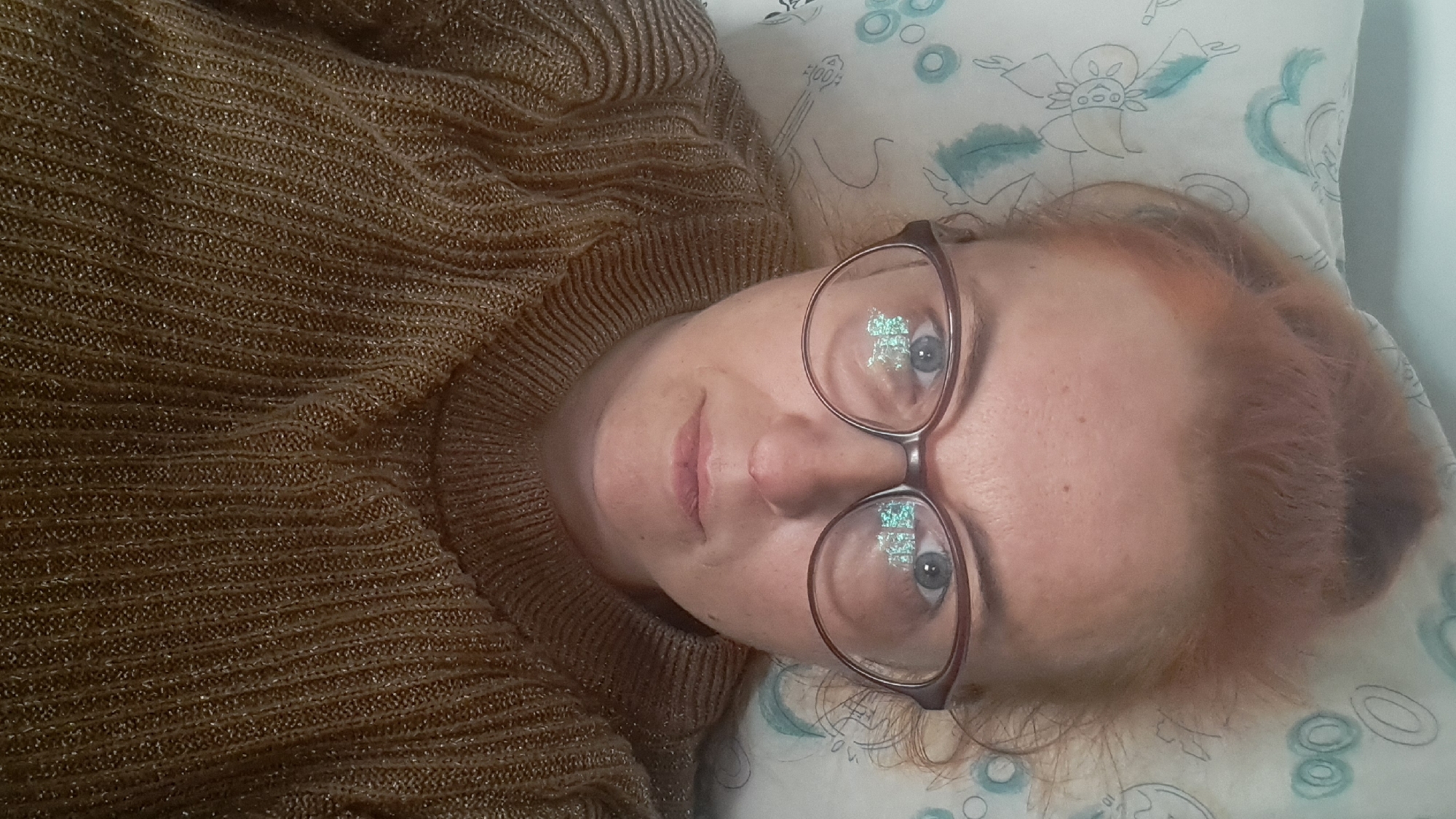 A close-up, overhead shot shows a woman lying down with her head on a pillow. She is wearing glasses with a reflective surface and a brown, sparkly sweater. The pillow has a light-colored pattern of stylized green leaves and flowers. The woman's hair is pulled up in a bun and appears to be reddish-brown. The reflection in her glasses shows a screen with a green, pixelated pattern, possibly from a computer or phone. The background is a plain, light color, likely a wall or ceiling. There is no indication of the location being in Riga, Latvia.