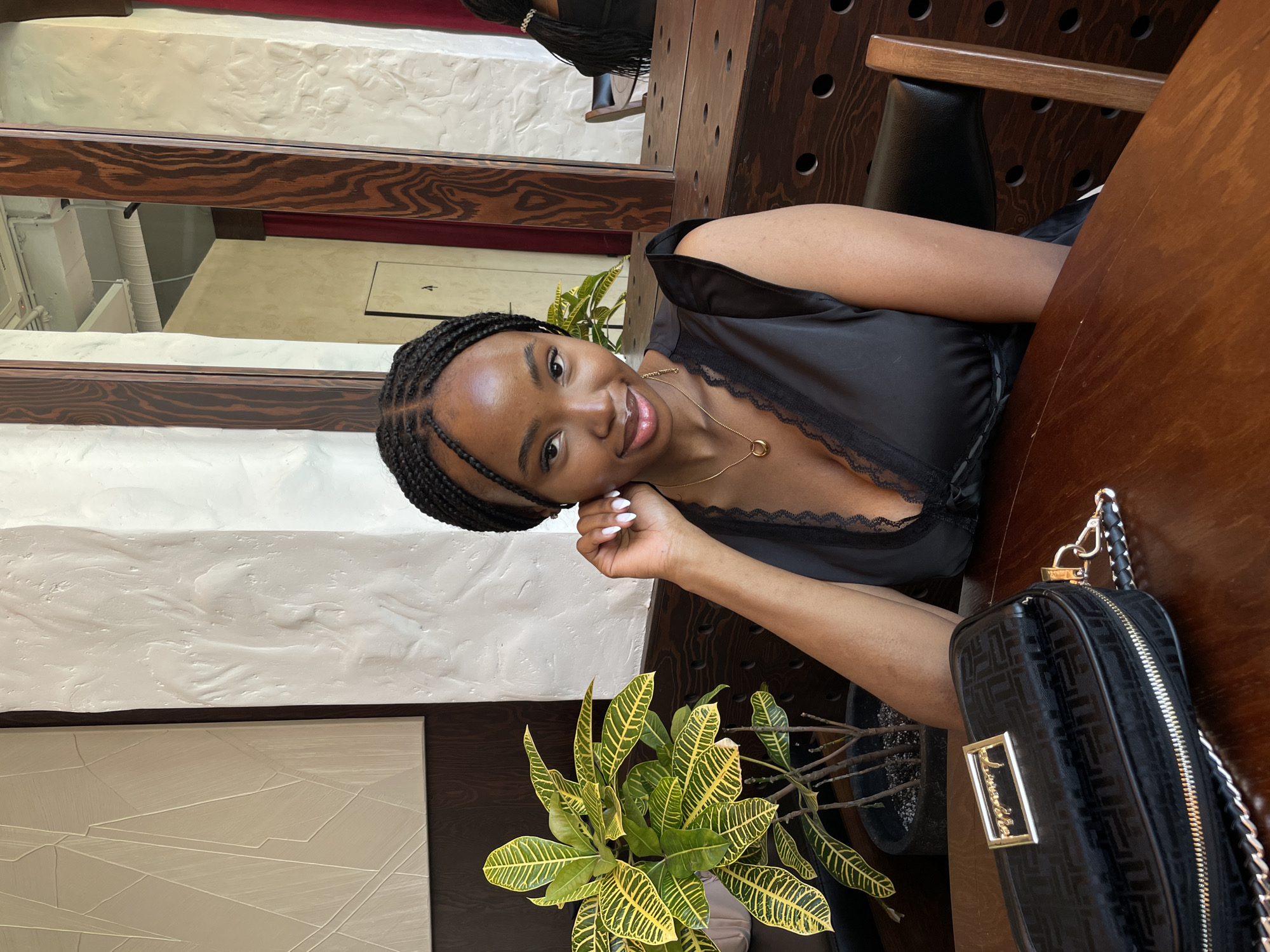 A young Black woman with braided hair and fair skin is sitting at a wooden table in a restaurant. She is wearing a black dress with lace trim and a gold necklace. In front of her, there is a black purse and a potted plant with green and yellow leaves. The background shows a textured white wall and a mirror reflecting another table and chairs. There is no indication that this scene is located in Kazan, Russia.