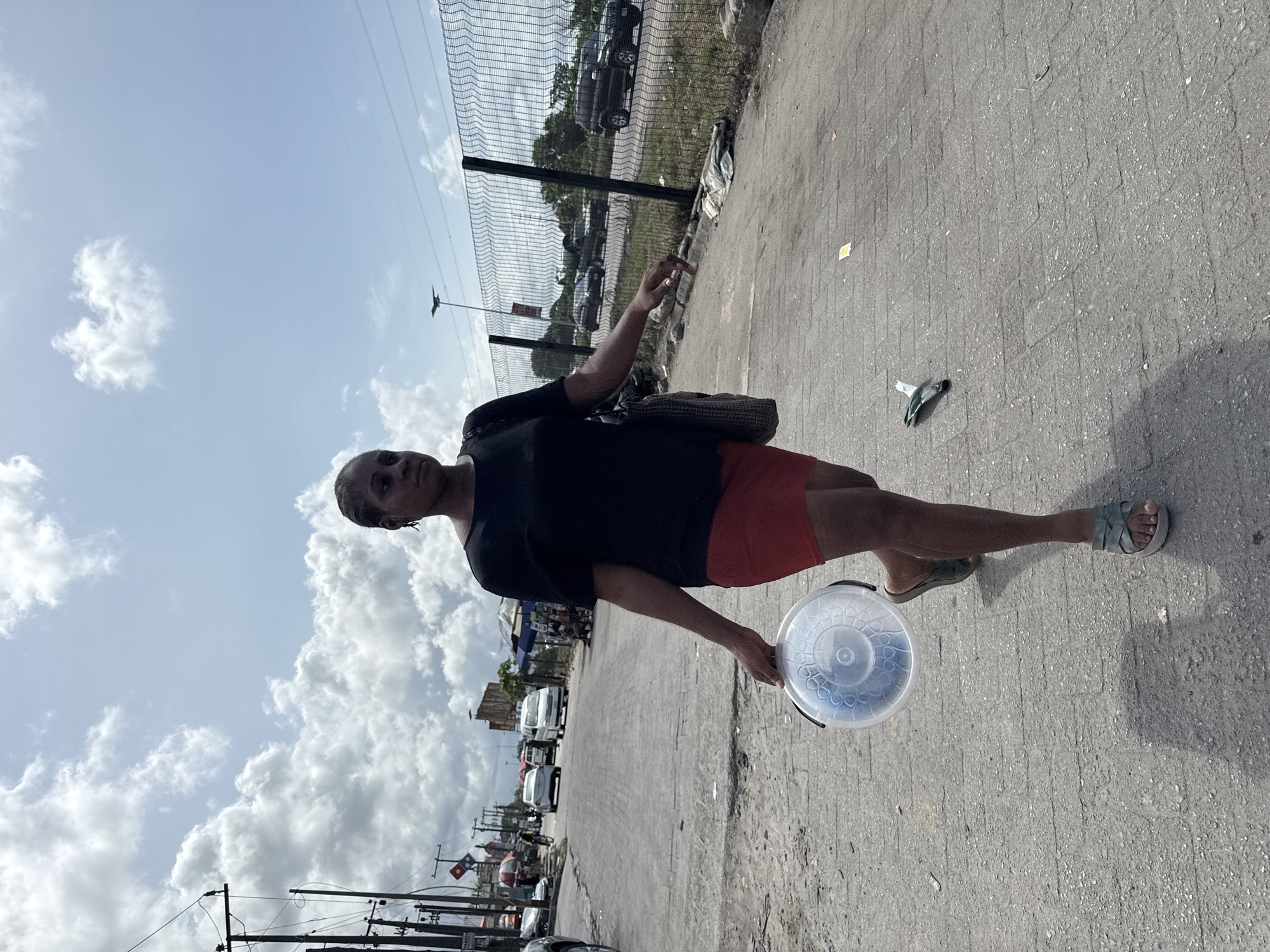A daylight street scene in Lagos, Nigeria, captures a woman standing by a busy roadside. The woman, who appears to be in her late teens or early twenties, has dark skin and short, braided hair. She wears a short-sleeved black top and red shorts, with light-colored sandals on her feet. In her right hand, she holds a translucent plastic bucket with a lid, featuring a circular, patterned design. A dark shoulder bag hangs from her left shoulder. Her expression is neutral as she looks towards the camera.
The ground she stands on is paved, appearing to be concrete or asphalt, with visible cracks and some small pieces of litter. To her left, a street or road stretches into the distance, lined with various cars and vans. Utility poles with power lines run alongside the road. Behind the woman, a tall chain-link fence separates the paved area from a grassy or dirt patch where more vehicles are parked or visible, along with distant trees. The sky above is bright blue with scattered white clouds, indicating sunny weather. Strong sunlight casts distinct shadows on the ground, suggesting it is midday or early afternoon. No legible text is clearly visible.
