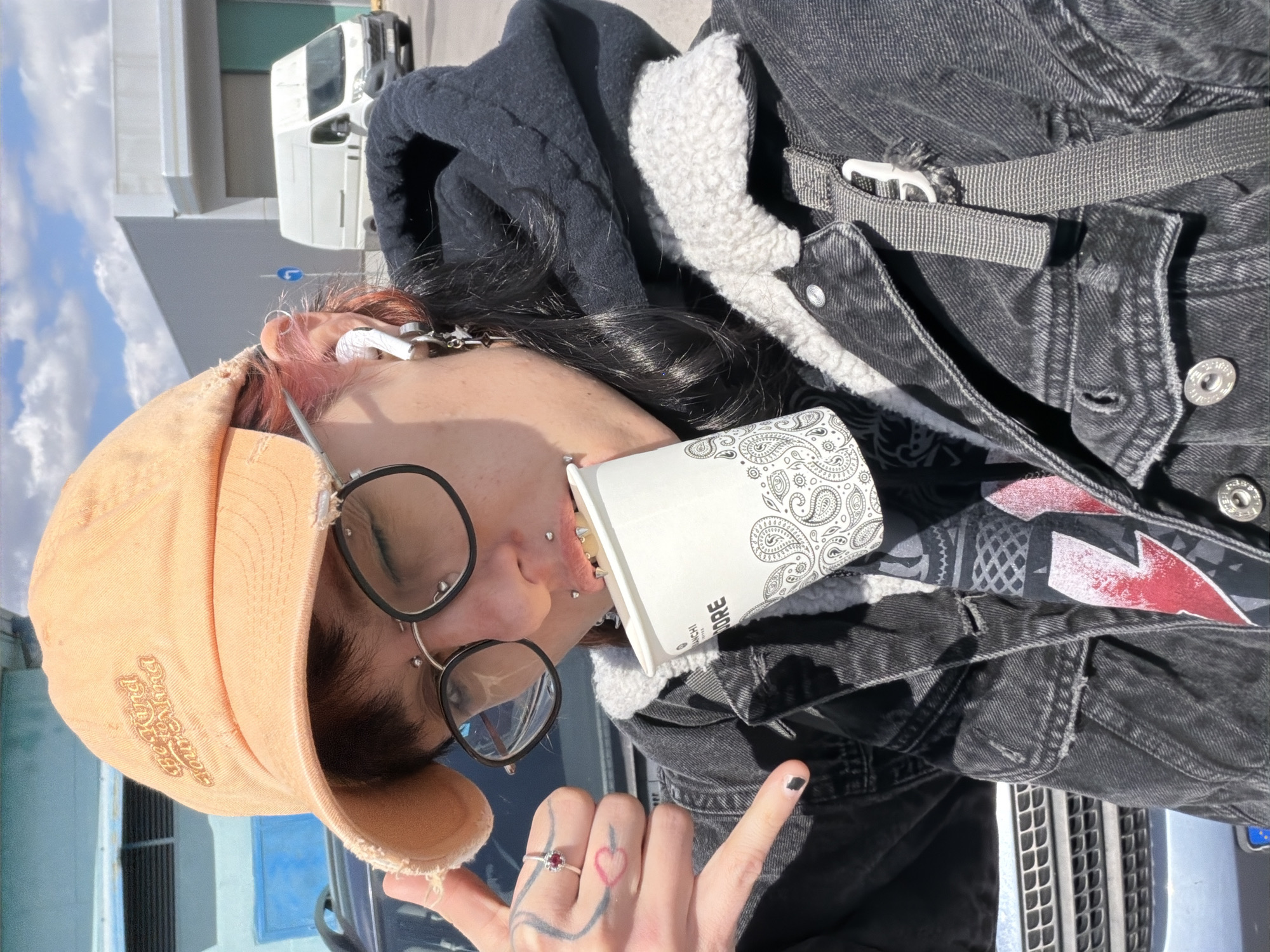 A person is holding a coffee cup and wearing a black jacket, peach cap, and glasses. The location appears to be an urban area, potentially Sofia, Bulgaria.
