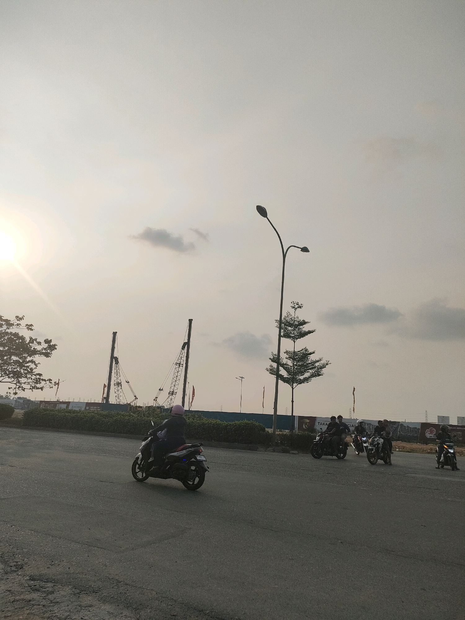 The image depicts a construction site adjacent to a roadway in Batam, Indonesia, under a hazy, overcast sky with the sun partially obscured. In the foreground, a motorcycle rider wearing a pink helmet is traveling on the asphalt road. Further down the road, several other motorcycles are visible, some with riders. The background features tall construction cranes, indicative of an ongoing development project. Along the edge of the road, there is a line of trees and bushes. Billboards are also visible behind the foliage, suggesting commercial or residential development. The overall scene conveys a sense of urban development and daily transit in Batam.