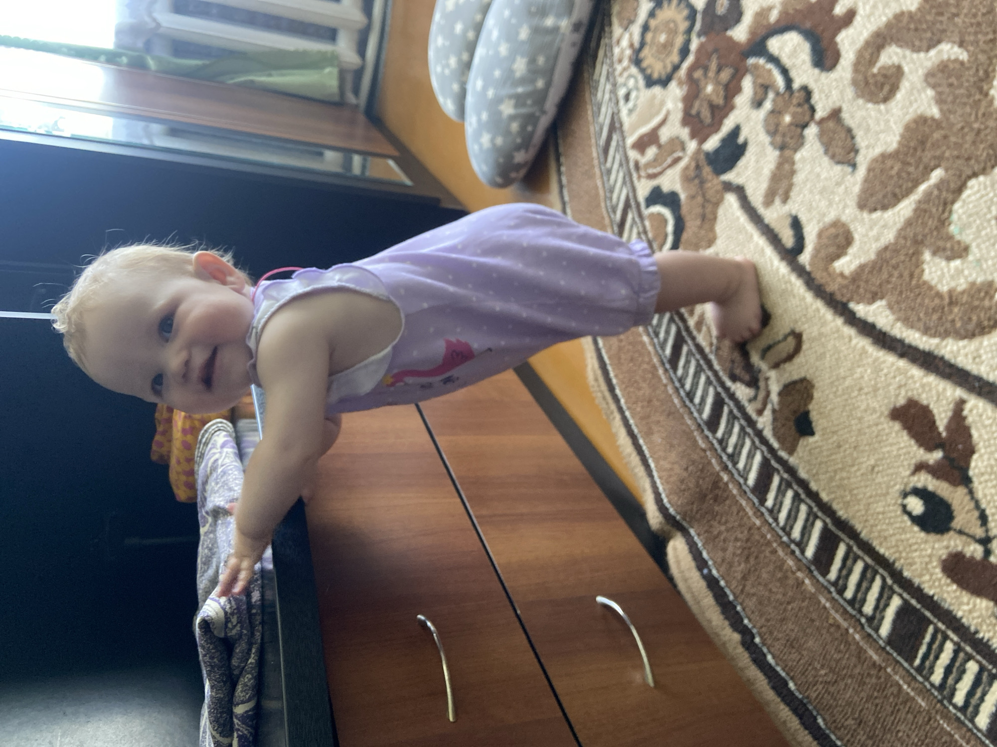 A baby in a purple romper is in a planking position between a dresser and a rug with an ornamental brown and beige design. The baby's hands rest on the top of the dresser, and her feet are planted on the edge of the rug. The background features a pillow, a window, and a partial view of a wooden floor. The location is Фоки, Russia.