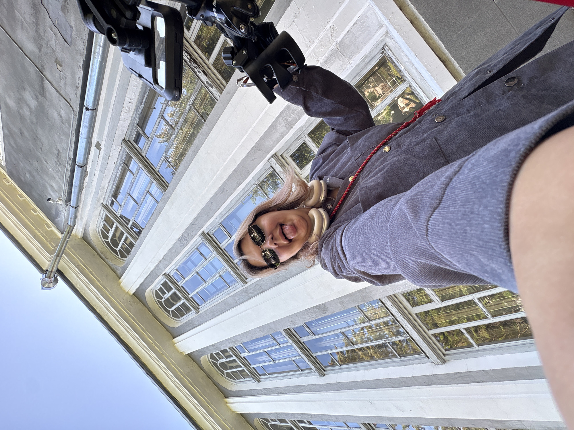 A low-angle shot captures a person taking a selfie with a camera mounted on a device. The person, wearing sunglasses and a grey jacket, sticks their tongue out at the camera. They have blonde hair and large headphones around their neck. Behind them, a grand building with tall windows and ornate architectural details is visible. The sky above is clear blue. The scene appears to be outdoors, possibly in a historical or institutional setting. No specific details in the image suggest the location is Birobidzhan, Russia.