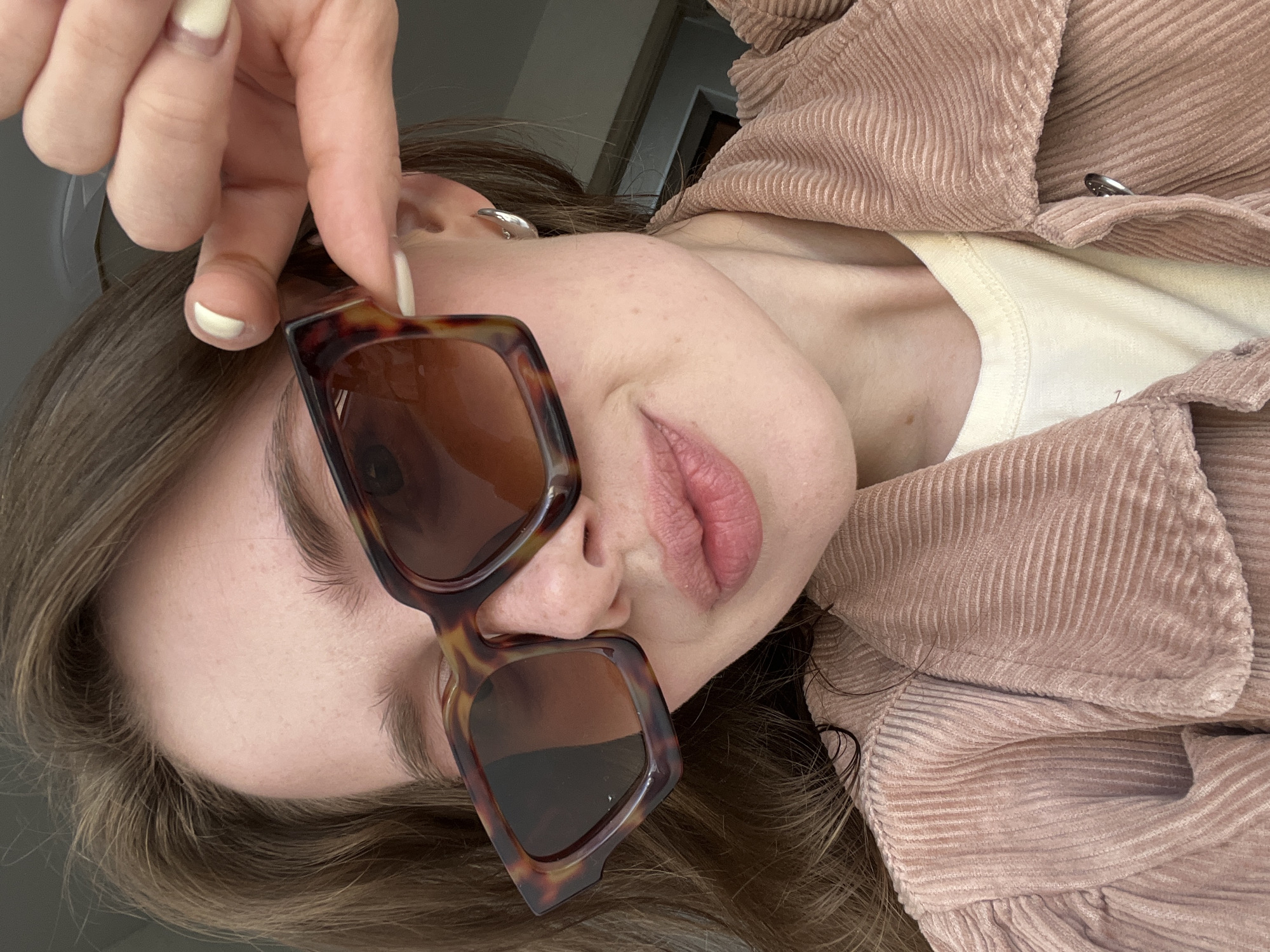 This close-up, angled portrait features a young adult, likely female, captured indoors in Chaikovsky, Russia. The subject's face is presented at an unusual angle, rotated approximately 90 degrees counter-clockwise, showing her from the forehead to the chin. She has fair skin, brown hair, and her lips are slightly parted, revealing a natural pink hue.
A prominent object in the foreground is a pair of large, square-framed sunglasses with a distinctive tortoiseshell pattern and dark lenses. These glasses are partially worn, with one lens covering her eye area and the other resting on her cheek and nose. Her left hand, with light-colored (off-white/cream) manicured nails, is visible holding the upper part of the sunglasses, appearing to adjust them. A small, silver-toned hoop earring is visible in her left ear.
The subject is dressed in a cream or off-white t-shirt or top, layered under a dusty rose or light brown corduroy jacket, which has a noticeable ribbed texture. The background is indistinct, suggesting a plain indoor setting. The overall mood is casual and relaxed. No visible text is present in the image.