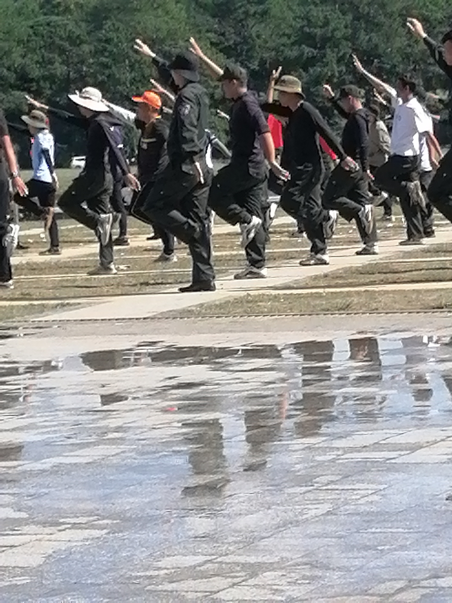 The image captures a group of people, primarily men and a few women, engaged in a synchronized physical activity, possibly a march or drill, in what appears to be an outdoor setting. The individuals are dressed in dark uniforms, with some wearing hats and others not. Their arms are raised in a uniform manner, suggesting a coordinated movement. The ground in the foreground is wet and reflective, indicating recent rain or a water feature, possibly a fountain or a puddle. In the background, lush green trees form a dense canopy under a bright sky. The scene suggests a structured event taking place in a park or an open space within a city. The specific location in Da Lat, Vietnam, is not explicitly identifiable from the visual cues in the image.