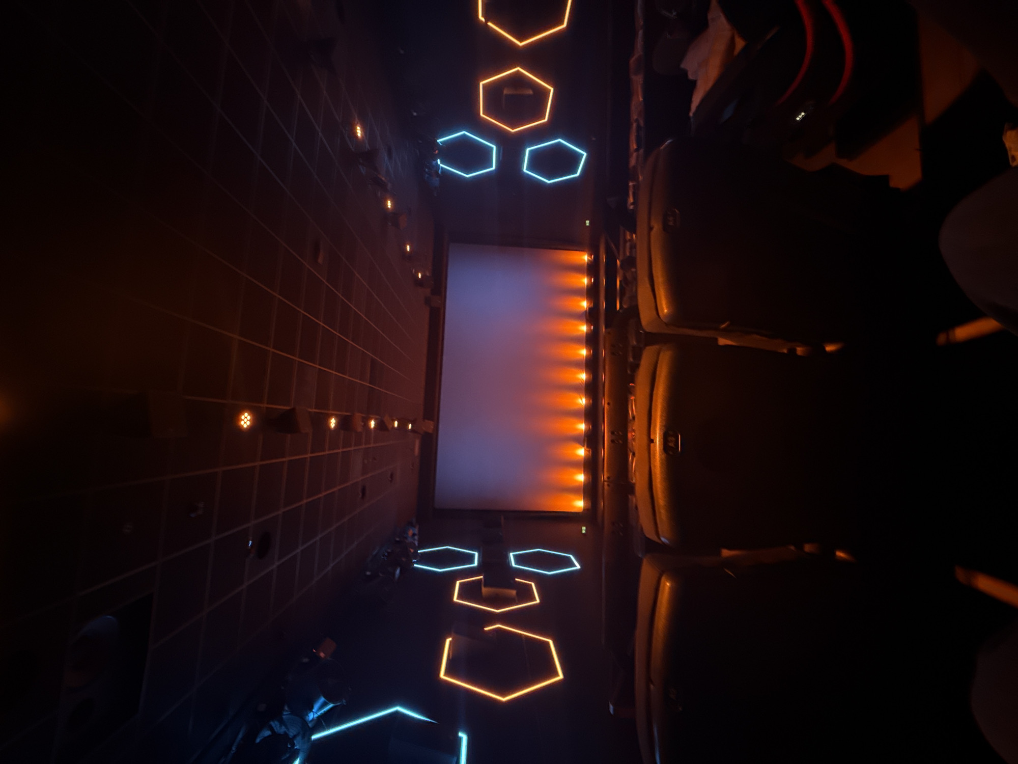 The image displays the interior of a dimly lit modern cinema auditorium in Kassel, Germany, captured from a rear seating perspective. A large, blank projection screen at the front exhibits a faint blue-white luminescence. Below the screen, a row of ten discrete, upward-facing lights casts an orange glow onto the lower wall. Both side walls feature decorative, neon-style hexagonal light fixtures; the left wall shows three orange and two blue hexagons, while the right wall displays three blue and two orange hexagons. The ceiling above consists of dark, grid-patterned panels with scattered small, warm-toned recessed lights. In the immediate foreground, the backs of several dark-colored cinema seats are visible, indicating the viewer's position. Further rows of seats extend towards the screen, gradually fading into the dim light. No individuals are clearly depicted, nor are any specific actions occurring.