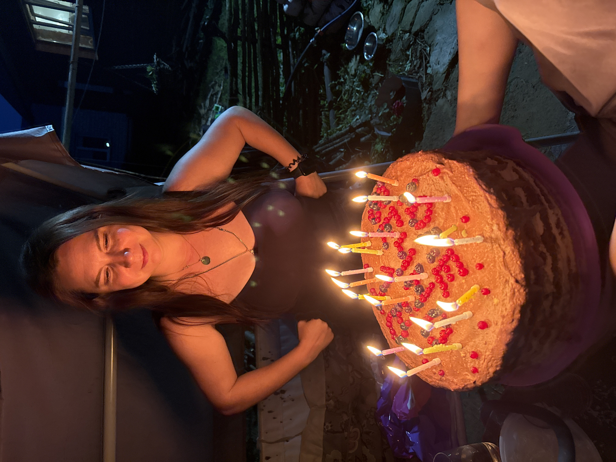 A medium shot captures a woman in a dark dress standing behind a large, multi-layered cake adorned with lit candles and berries. She is looking down at the cake with a slight smile. The cake is placed on a purple stand. The background suggests an outdoor setting at night, possibly a backyard or a rural area, with a dark wooden fence and a structure resembling a house with illuminated windows. The lighting is dim, with the primary light source coming from the candles on the cake and some ambient light illuminating the woman and the cake. There are hints of vegetation and rustic elements in the environment. The scene appears to be a birthday celebration.