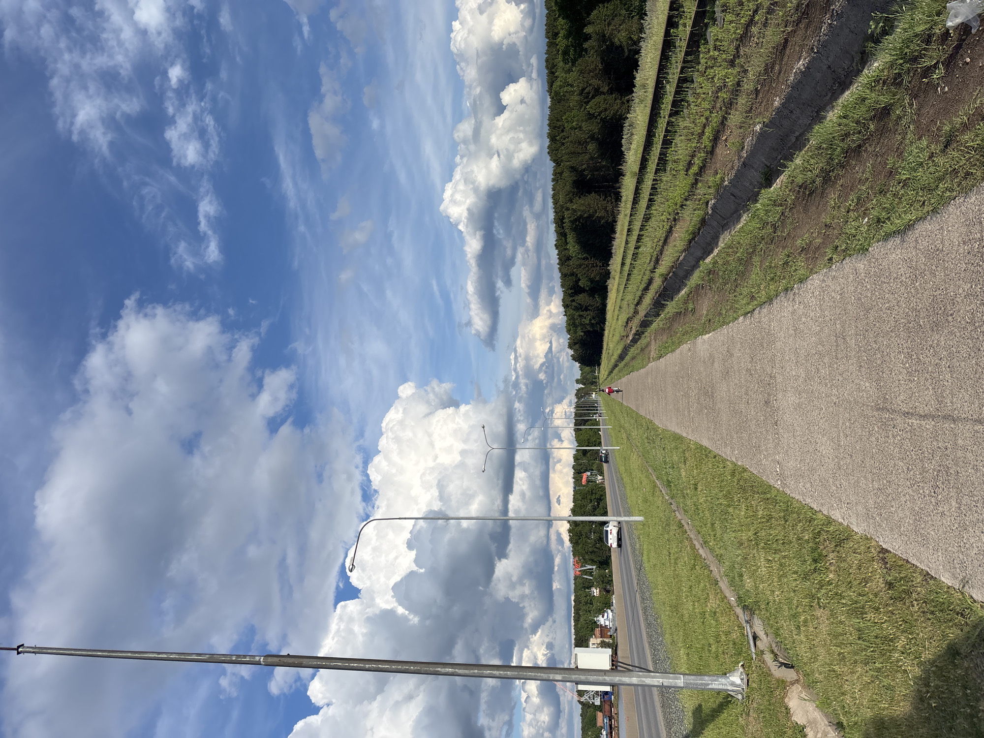The image shows an outdoor scene in what appears to be the city of Chaikovsky, Russia, based on the landscape and infrastructure. A long, concrete path stretches into the distance, flanked on one side by a grassy area and what looks like newly planted rows of vegetation or saplings. On the other side of the path, there's a road running parallel, with intermittent streetlights. Further beyond the road, there are buildings and trees, suggesting a residential area. A person is visible on the path, walking or running in the distance. The sky occupies a significant portion of the image, filled with billowing white and gray clouds against a blue backdrop.
