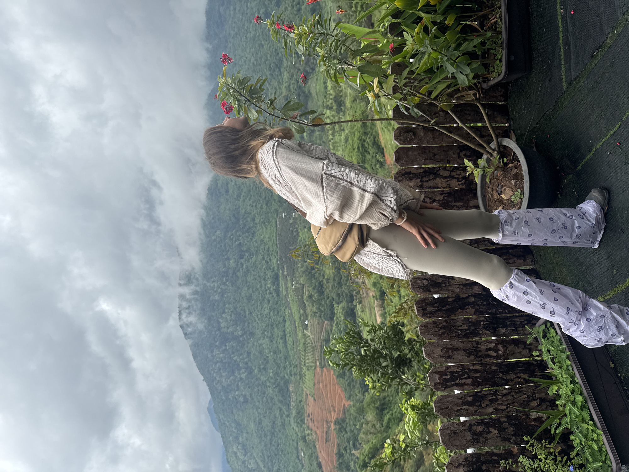 A young woman with long blonde hair, wearing a white patterned blouse, beige leggings, and white patterned leg coverings over her boots, is shown from the side, leaning forward to smell a small plant with red flowers. She has a light brown fanny pack around her waist. The scene is set on a balcony or viewing platform with a rustic wooden fence, overlooking a lush green mountainous landscape. In the background, terraced hills with reddish-brown soil are visible, along with patches of trees and some small white structures. The sky is overcast with heavy clouds, partially obscuring the tops of the distant mountains. The location is identified as La Dạ, Vietnam.
