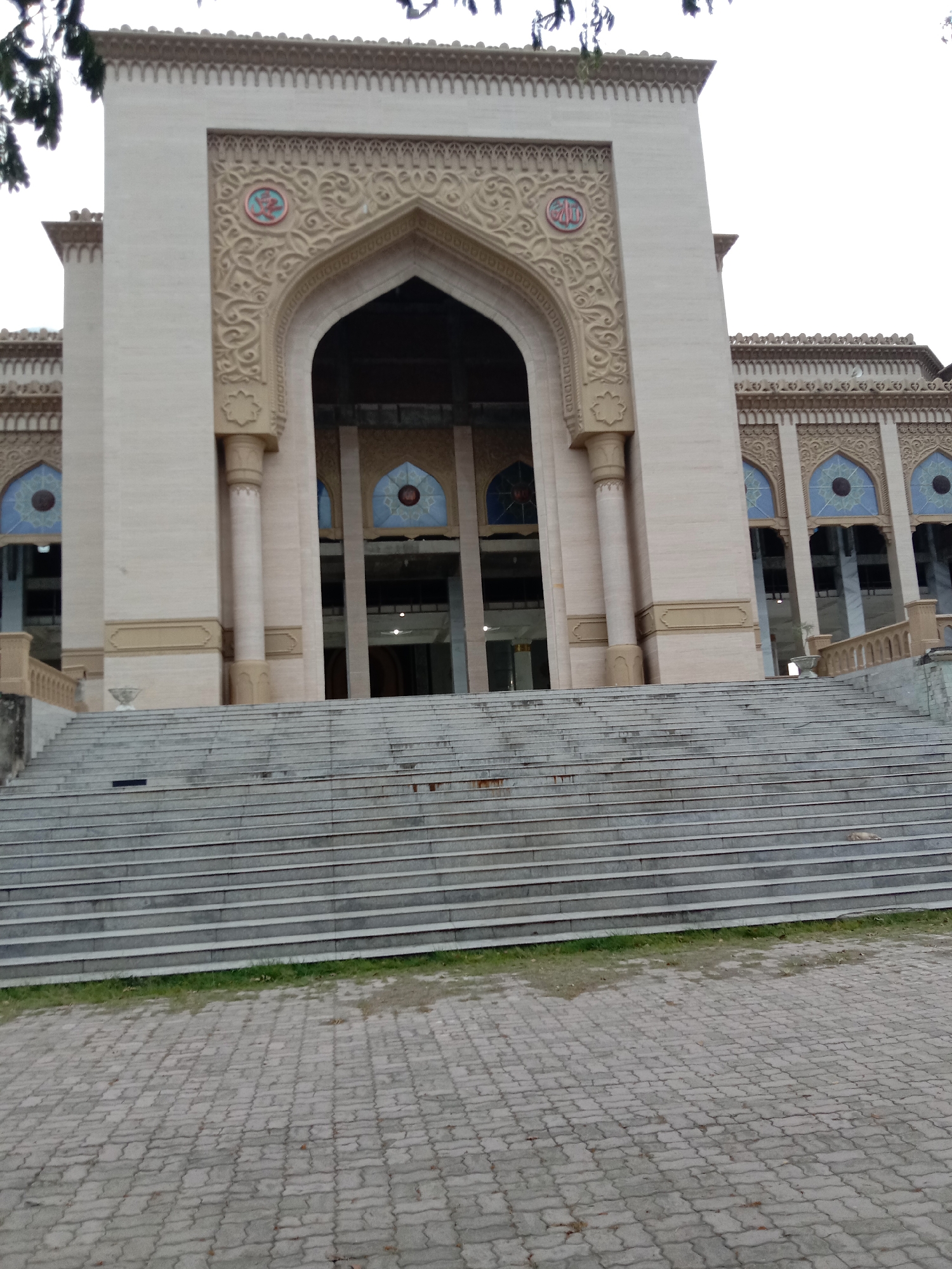 The image displays the grand front entrance of a large building, likely a mosque, situated in Lhokseumawe, Indonesia. The central feature is a wide, imposing staircase, constructed from numerous grey stone or concrete steps, leading up to the main edifice.
The building itself is light-colored, possibly stone or marble, and exhibits ornate Islamic architectural characteristics. A prominent, tall arched entryway forms the main portal, flanked by sturdy columns. The facade surrounding this arch is richly decorated with intricate, symmetrical floral and geometric bas-relief carvings. Above the main arch, two circular medallions are embedded in the decorative stonework. The medallion on the left clearly contains stylized Arabic calligraphy, identifiable as the word "Allah" (الله), while the right medallion also features similar religious script.
On either side of the central entrance, the building extends with further architectural details, including additional arched windows set into recessed sections, also adorned with decorative blue glass elements and similar carvings above them. Low, decorative balustrades flank the base of the building where the staircase begins.
In the foreground, the ground is paved with interlocking grey concrete pavers, separated from the bottom step by a narrow strip of green grass. Dark foliage from a nearby tree is visible in the upper left corner of the frame. The lighting suggests an overcast daytime, with soft, diffused illumination and no harsh shadows. There are no people visible, and no specific activity or event is taking place, presenting a serene and majestic scene.