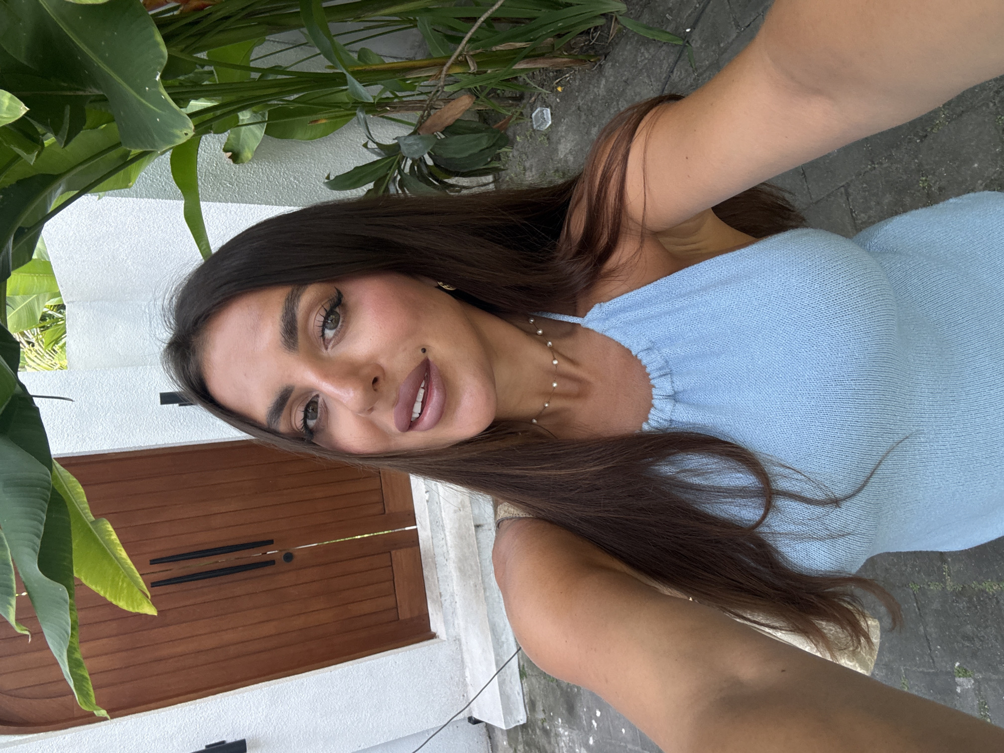 A selfie captures a woman with brown hair, light skin, and defined features. She is wearing a light blue dress and a delicate gold necklace. Her face is angled slightly, with a visible mole on her right cheek. The background reveals a modern architectural structure with a wooden door, white walls, and lush green tropical foliage. The scene suggests a location in North Kuta, Indonesia, given the tropical vegetation and architectural style.