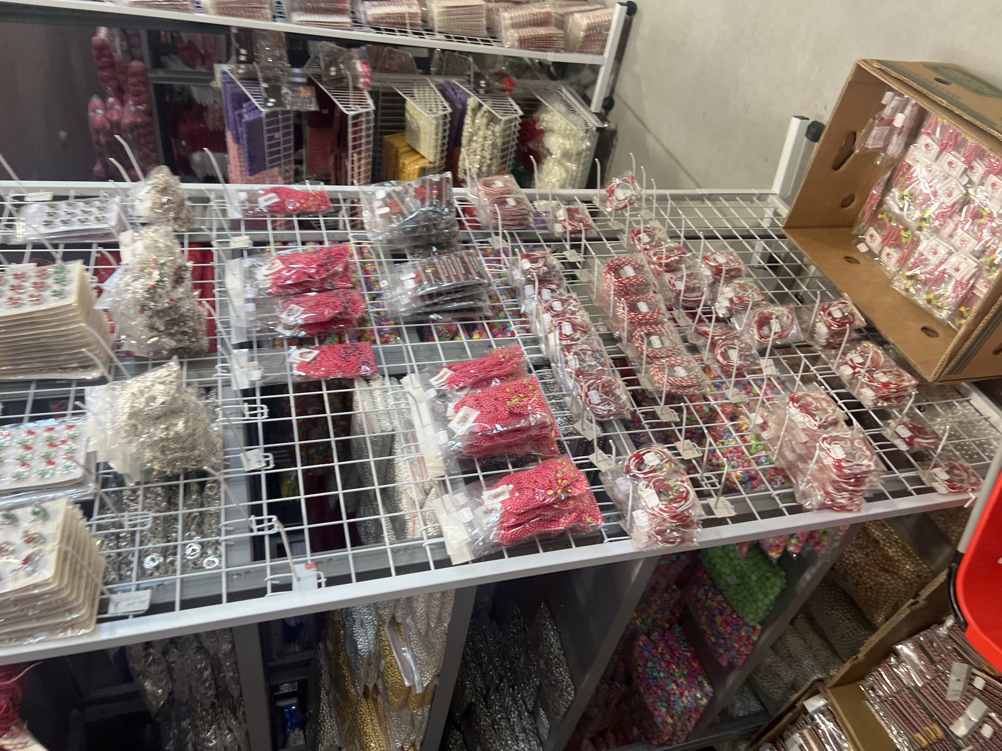 The image shows a retail setting with multiple shelves displaying various packaged items. Predominantly, these items appear to be craft or hobby related, possibly for jewelry making or decorative purposes, including beads and small decorative pieces.
The items are organized and presented in a grid-like arrangement on white metal shelving. The shelves are stocked densely with different colors and types of packaged goods. A cardboard box on the right side is filled with packets of similar items.
The environment looks to be the interior of a retail or wholesale store specializing in craft supplies. No people are visible. The background shows additional shelves with similar items, indicating a larger store layout.