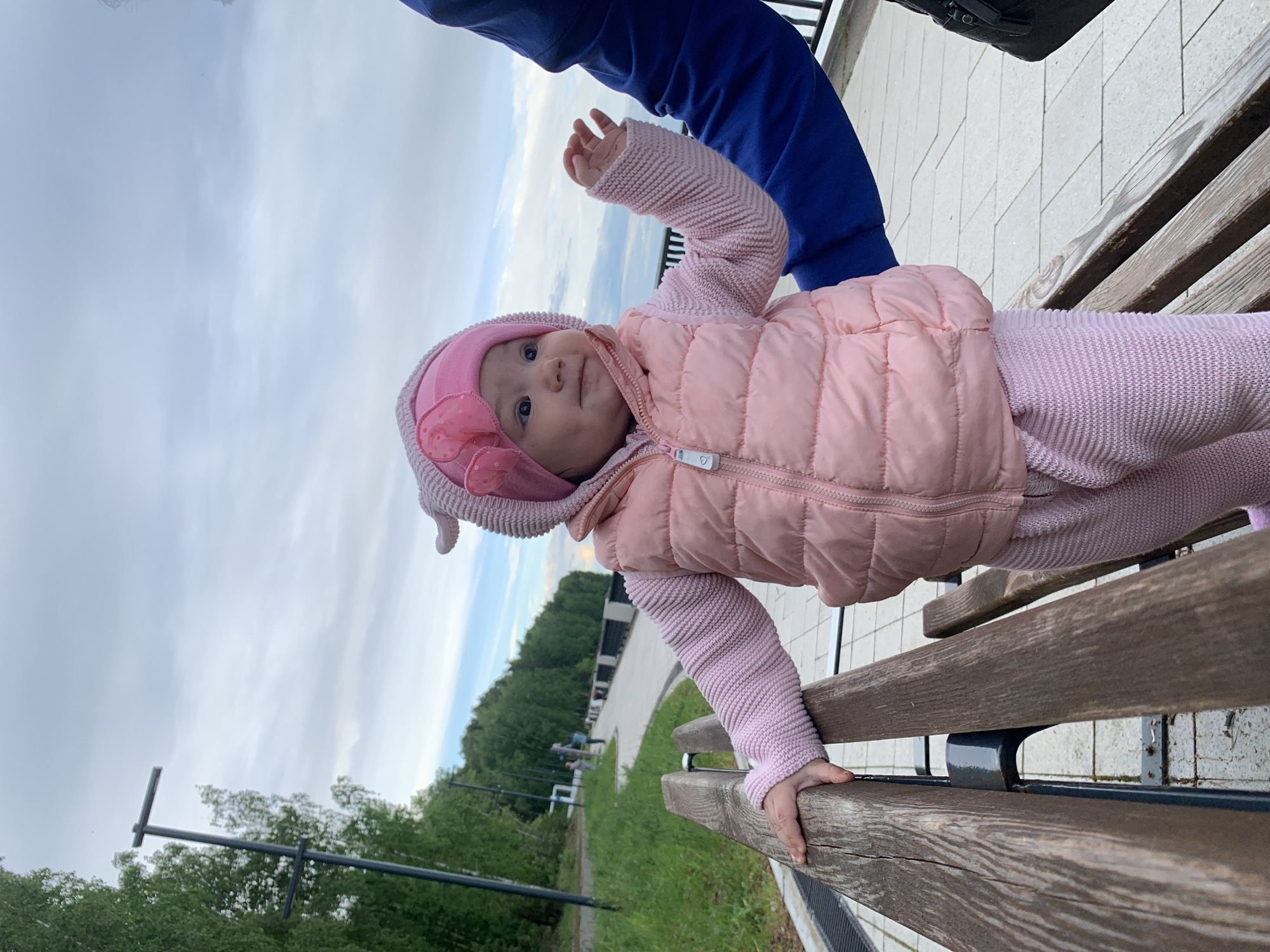 A baby wearing a pink winter hat and vest is standing on a wooden bench. A person wearing a blue jacket is standing behind the baby. The setting is outdoors, with green grass and trees visible in the background. There is a paved path and street lights in the distance. The image was taken in Chaikovsky, Russia.