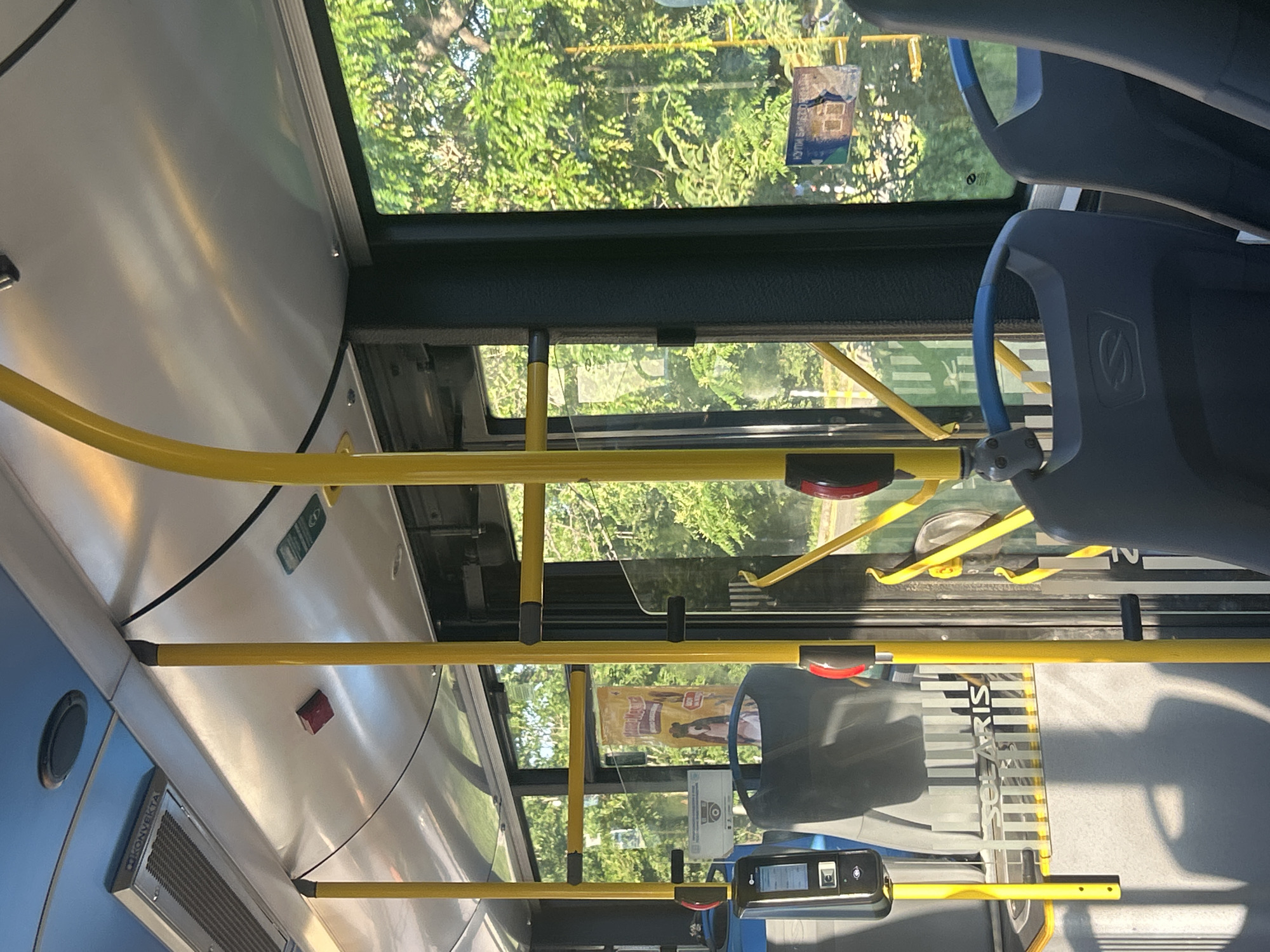 The image shows the interior of a public bus with yellow poles and blue seats. Sunlight streams in from the windows, illuminating the reflective ceiling and casting shadows on the floor. Outside the windows, green trees are visible. A ticket validator is mounted on a pole near the front of the bus. The overall impression is a bright, modern bus interior on a sunny day.