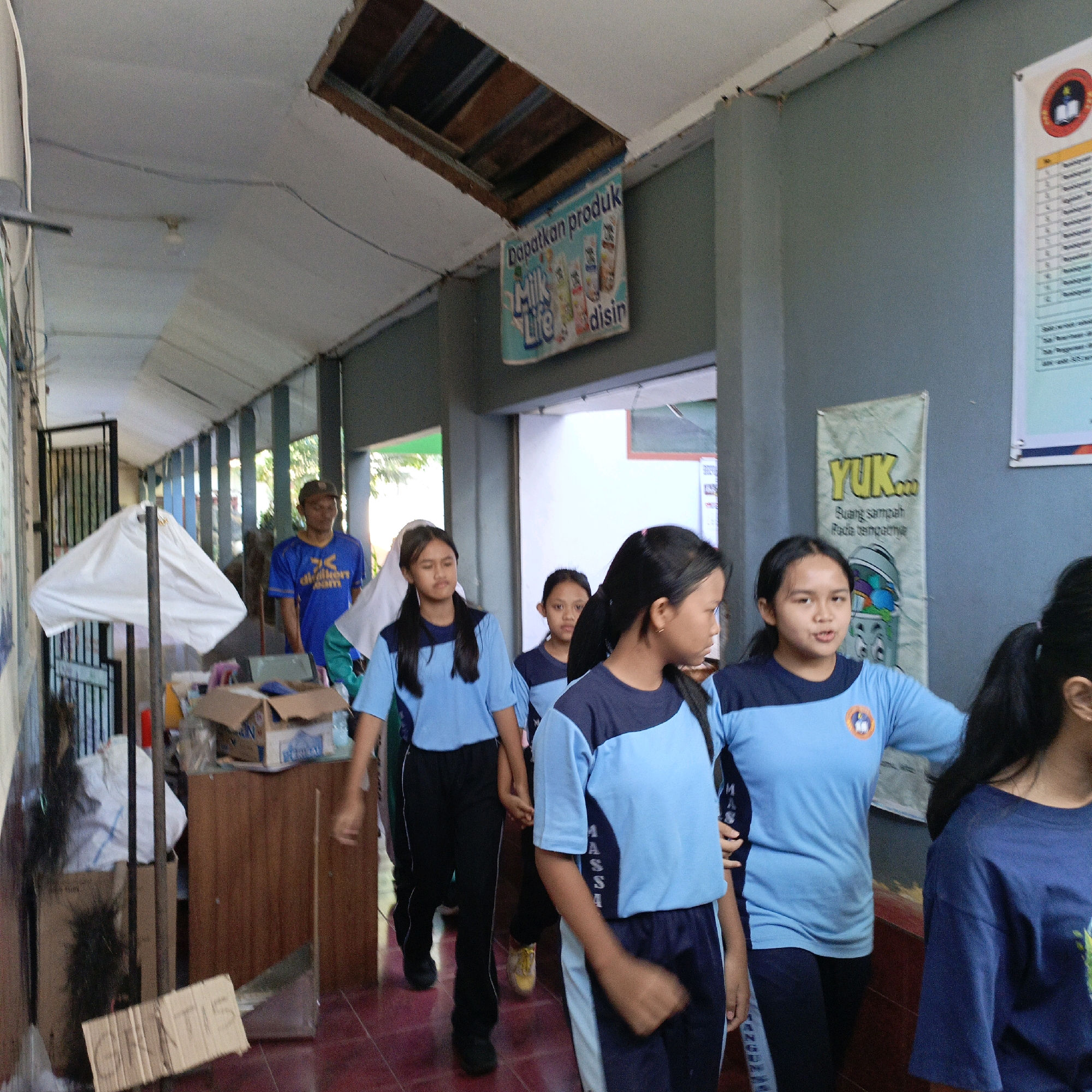 In Salatiga, Indonesia, a group of young females in light blue and navy blue uniforms walk down a covered walkway with a damaged ceiling. A man in a blue t-shirt and cap stands in the background. On the wall, a poster reads "YUK... Buang sampah Pada tempatnya," which translates to "LET'S... Throw trash in its place." Another poster advertises "Milk Life" products. Cardboard boxes and other items are stored along the walkway.