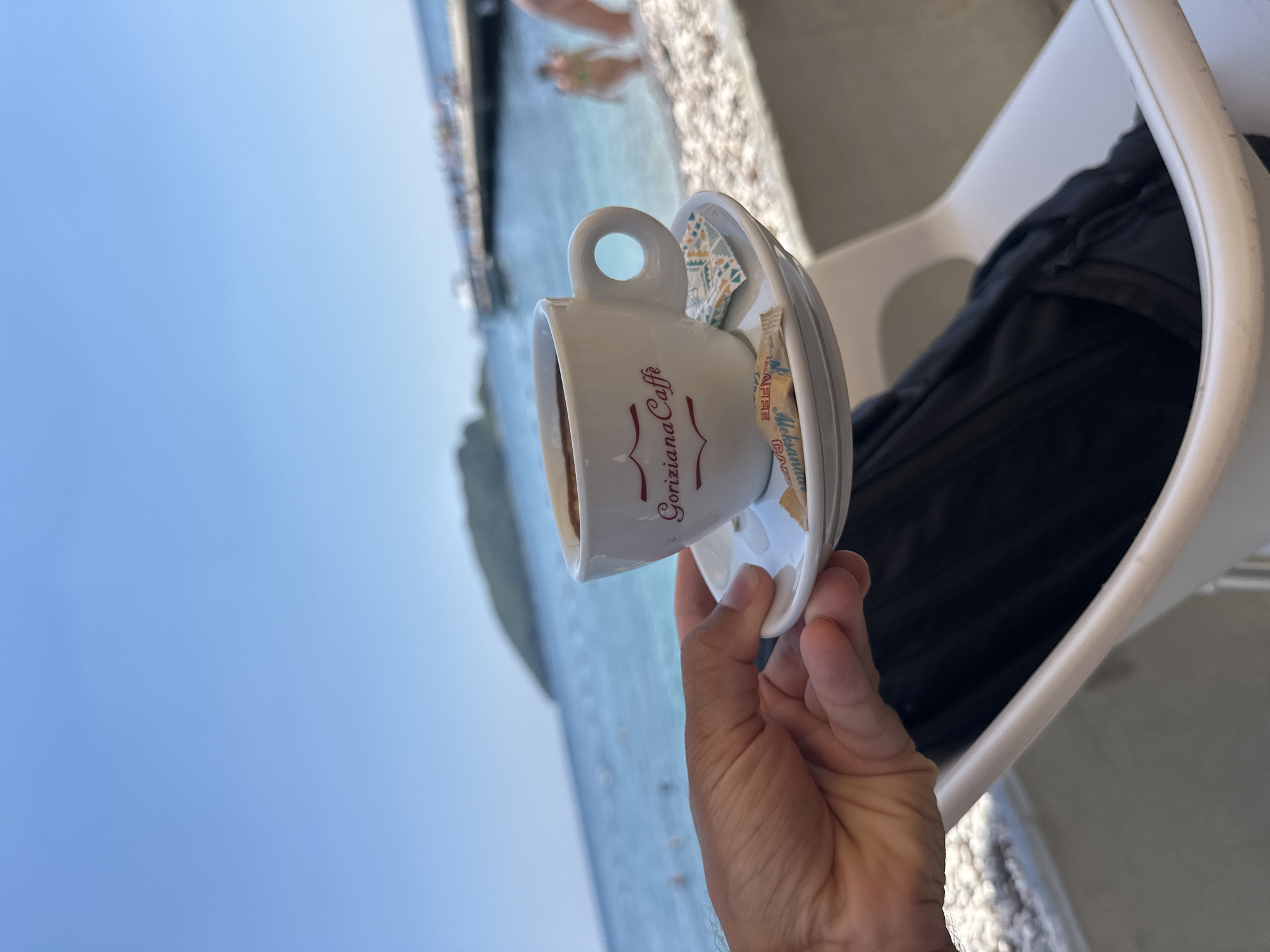 An outdoor, close-up shot captures a hand holding a white ceramic cup of dark coffee on a matching saucer. The cup has "Goriziana Caffè" written on it in red script. A small packet of sugar and a decorative napkin are also on the saucer. In the background, a bright blue ocean meets a pebbly beach, with a distant green-covered hill and a pier with people visible. The scene suggests a cafe setting by the sea in Budva, Montenegro, likely during daylight hours given the bright sky and sunlight.