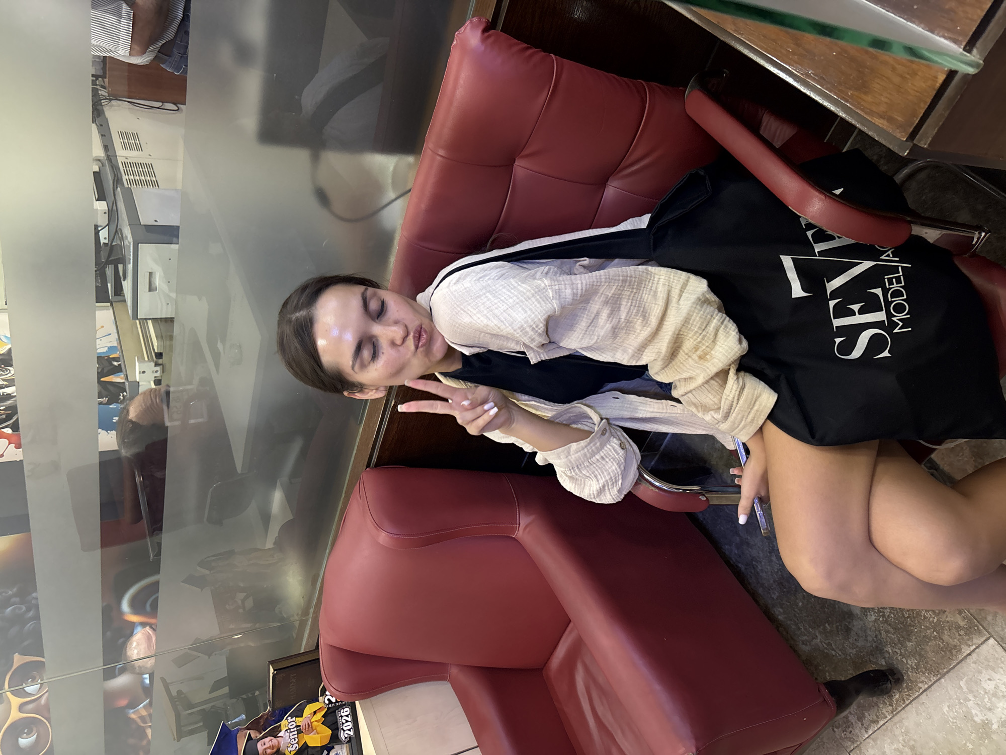 A young woman with brown hair is sitting in a red leather armchair, puckering her lips and holding up two fingers in a peace sign. She is wearing a black t-shirt, a light-colored open cardigan, and a black tote bag with the logo "SEVEN MODEL AGENCY" printed on it. Her legs are crossed. The background shows a frosted glass partition with reflections of an office or waiting area, including what appears to be office equipment and a graduation-themed poster. The flooring is tiled. The scene is indoors, likely in a commercial establishment. There is no specific indication of the city of Amman, Jordan, in the image itself.