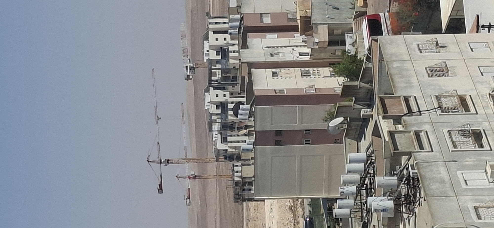 The image depicts an aerial view of a construction site and residential buildings in Dimona, Israel, under a clear, hazy sky. Multiple construction cranes are visible in the background, suggesting ongoing development. The foreground and midground are dominated by apartment buildings, some of which appear to be under construction or recently completed, characterized by their modern architectural style with light-colored facades and geometric designs.
Several rooftop water tanks, a common feature in Israeli residential buildings, are prominent on the roofs of the apartment blocks. Some buildings have satellite dishes and air conditioning units mounted on their exteriors. The landscape surrounding the urban development appears arid and undeveloped, typical of the Negev Desert region where Dimona is located. In the lower right section of the image, a red and white vehicle, possibly a bus or service vehicle, is partially visible. No individuals are discernible in the scene. The overall impression is one of a developing urban area in a desert environment.