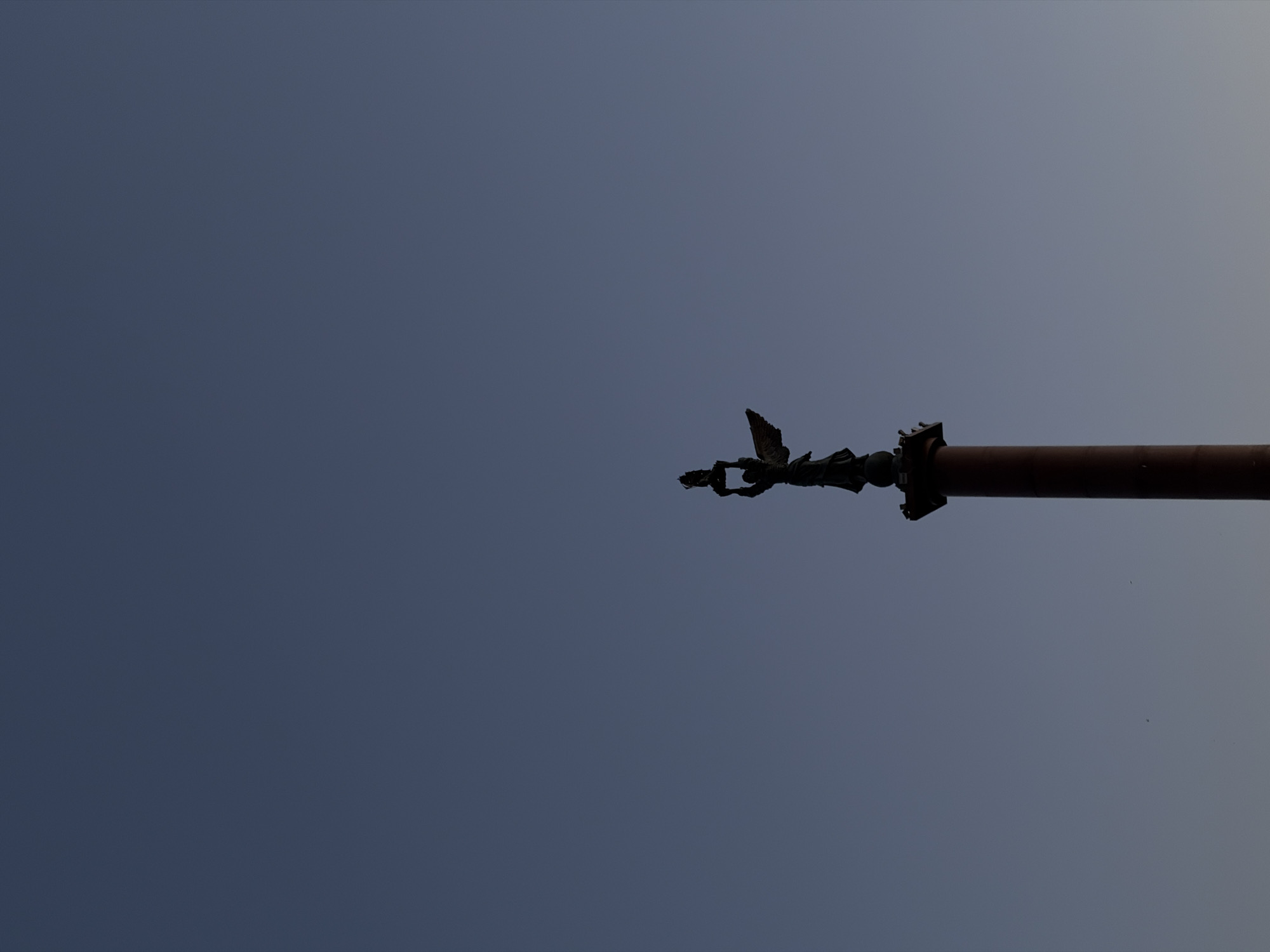 The image, taken from a low angle looking upwards, shows the top portion of a metal pole extending horizontally across the frame against a pale blue sky. The pole is a reddish-brown color and has some decorative metalwork at its end, which appears somewhat weathered or rusted. The location is undetermined, as there are no discernible landmarks.