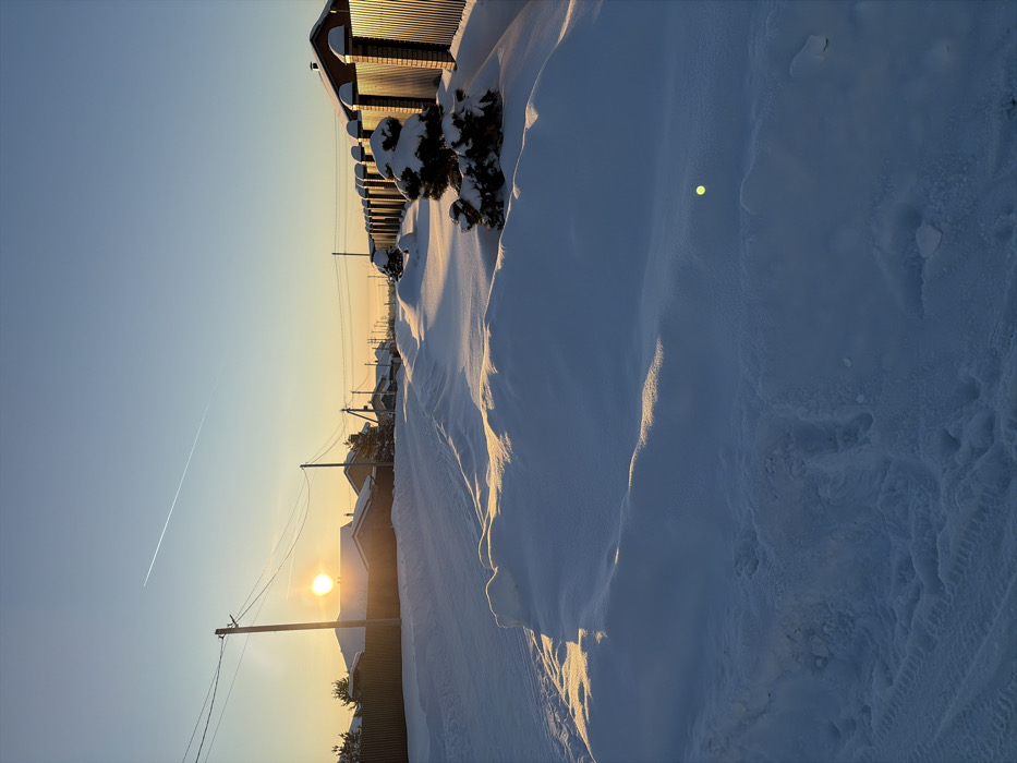 The image displays a snow-covered residential street in Stary Chultem, Russia, at sunrise or sunset. The sun is low on the left horizon, partially obscured by a distant roof, casting warm, golden light across extensive snowdrifts. These drifts line both sides of the unpaved or snow-covered road, where faint vehicle tire tracks are visible in the snow on the left. Utility poles and overhead lines run along the left side, adjacent to fences and residential buildings. On the right, a long corrugated metal fence with brick pillars parallels the street, interspersed with several small, snow-laden evergreen bushes. Distant houses line both sides of the road. A single aircraft contrail marks the clear blue sky. A small, circular green lens flare is visible in the lower right quadrant of the frame. No individuals are present.