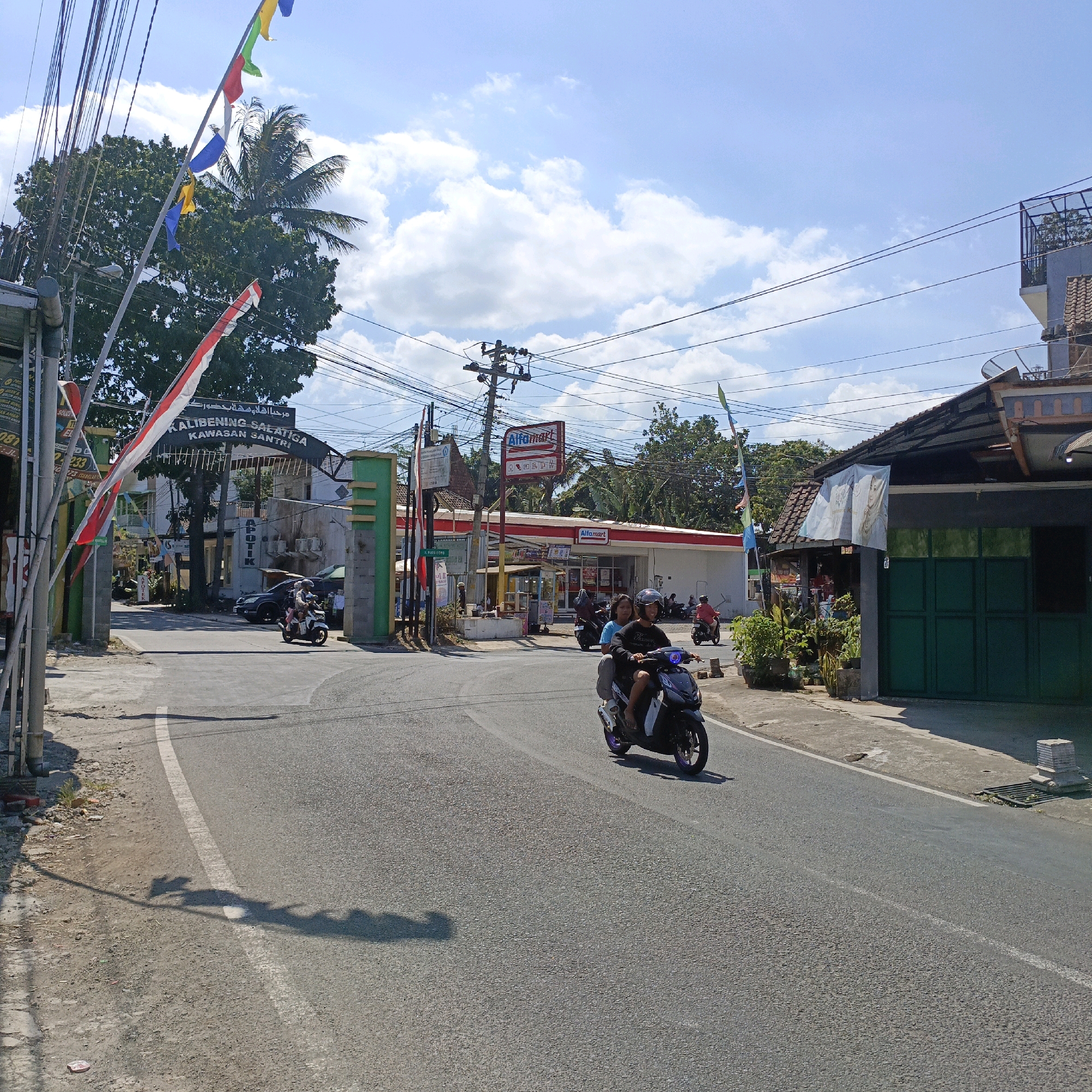 The image shows a street scene in Salatiga, Indonesia, under a bright blue sky with scattered clouds. In the foreground, a paved road curves to the right. Two people are riding on a dark-colored scooter, with the passenger in the back wearing a blue shirt and the driver in a black shirt and helmet. The scooter is heading away from the viewer. The road is marked with a white line.
On the left side of the road, there is a gateway with green pillars and a sign above it that reads "KALIBENING SALATIGA KAWASAN SANTRI". Indonesian flags, red and white, are displayed on poles. A large tree with dark green foliage is behind the gateway, with palm trees visible further in the background. Electrical wires are strung across the upper part of the image.
Further down the road on the right, there is an Alfamart store with its red and white signage. Several other motorbikes are parked or being ridden near the store. A building with a dark green door and some potted plants is also visible on the right. The overall impression is of a sunny day in a busy, somewhat cluttered urban environment.