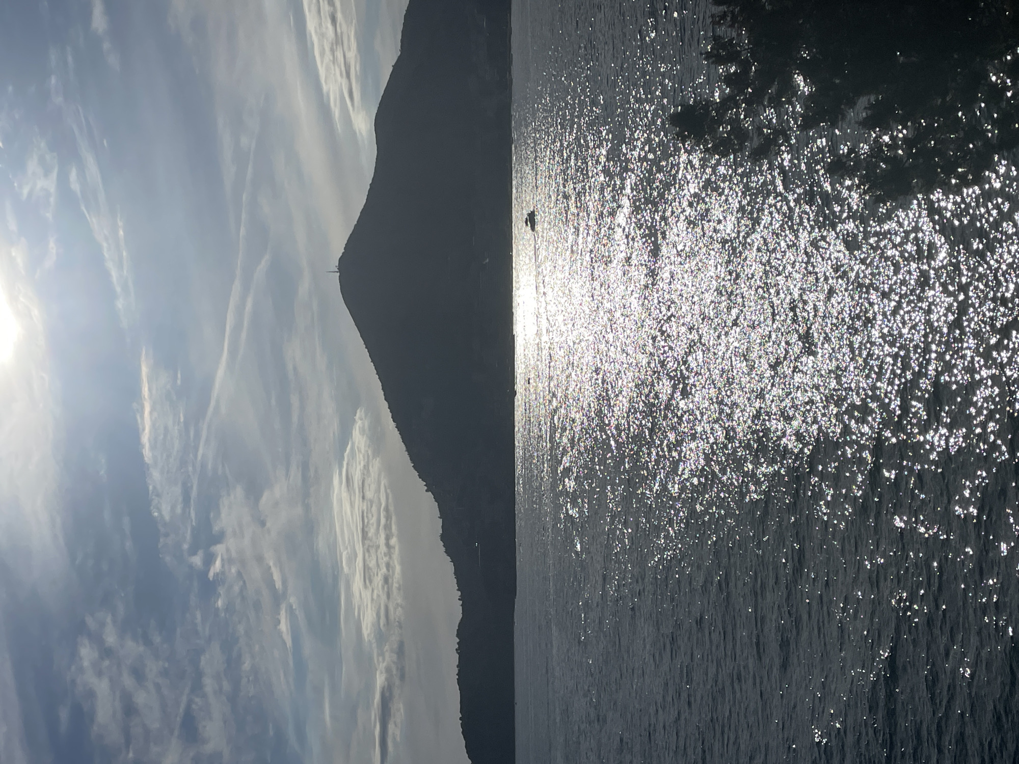 A silhouette of a conical mountain with a tower at its peak dominates the horizon, set against a bright, hazy sky with scattered clouds. Below the mountain, a dark, undulating coastline is visible. A wide expanse of water fills the foreground, reflecting the sunlight in a shimmering, scattered pattern. A small boat is visible in the distance, cutting across the water. The scene is likely Budva, Montenegro, looking towards Sveti Stefan or a similar coastal mountain. The light suggests it is either early morning or late afternoon.