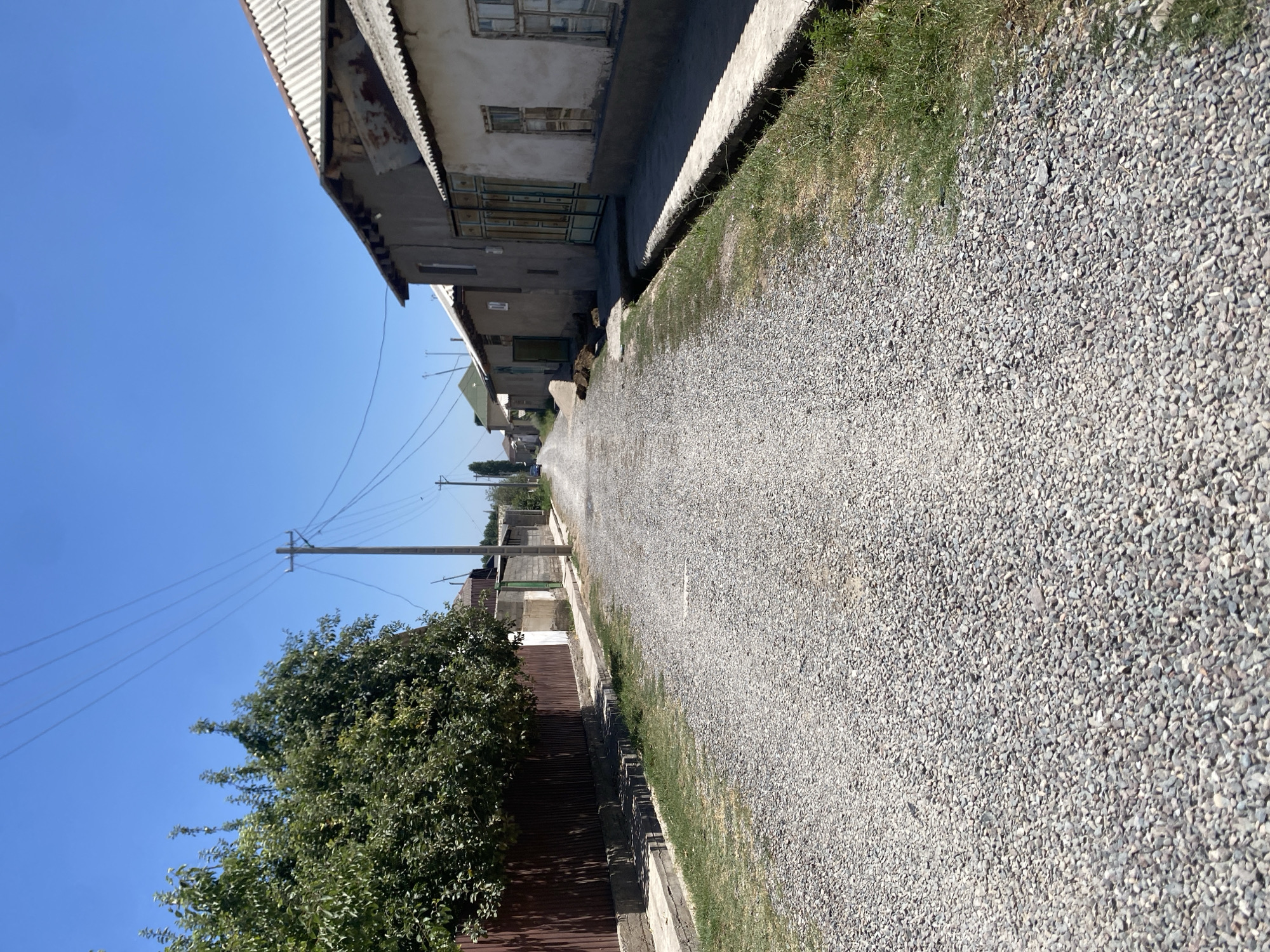 The image depicts a rural street in the city of Боғистон, Tajikistan. The street is unpaved and covered with gravel, running between a row of houses on the right and a fence and trees on the left. The houses appear to be constructed from stucco and have corrugated metal roofs. Power lines are visible overhead, supported by wooden poles. The sky is clear and blue, indicating a sunny day. There are no people or vehicles visible in the immediate scene, though a car can be seen in the distance down the road. The environment is typical of a village or suburban area with modest housing and infrastructure.