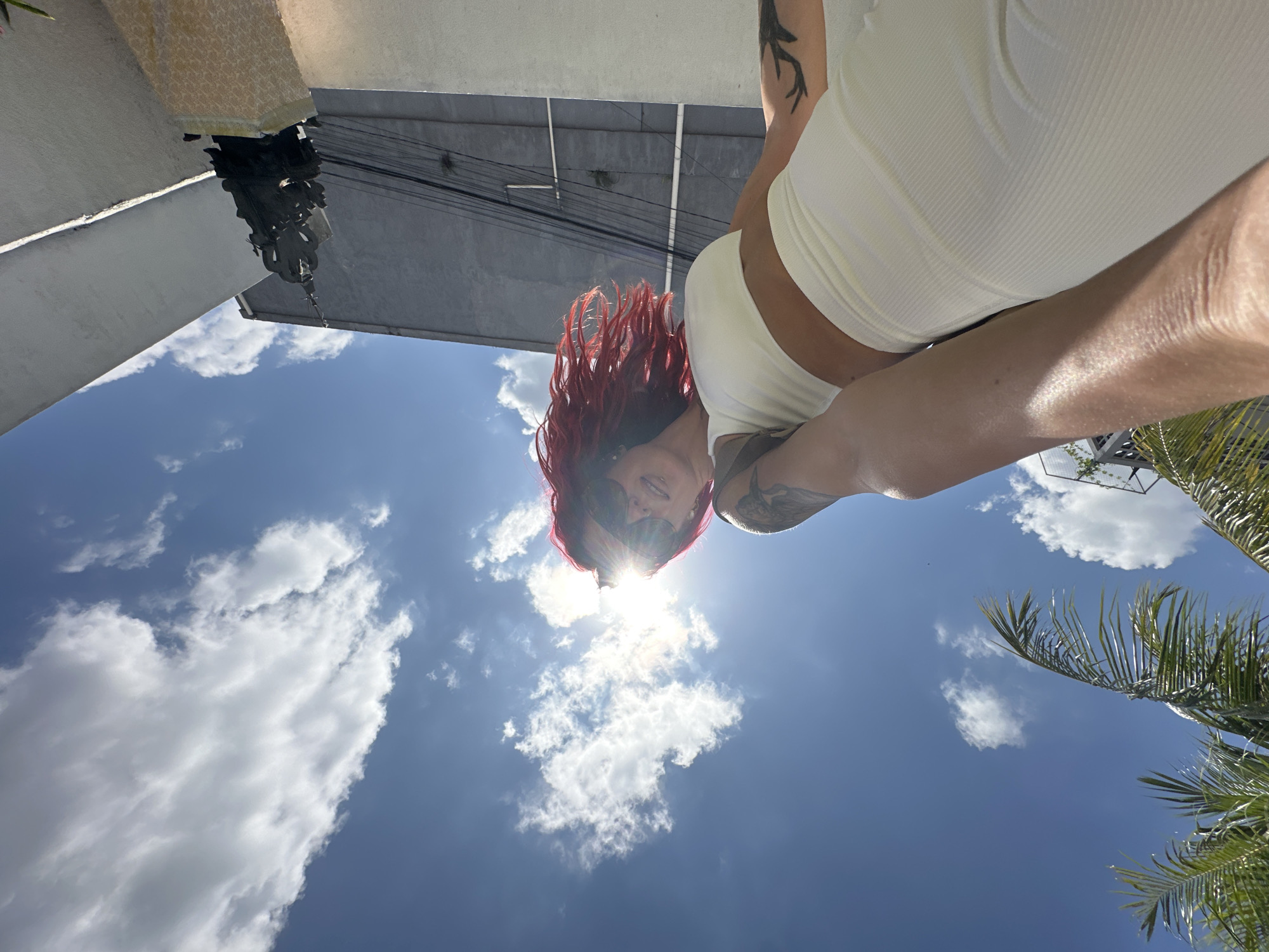 A low-angle, wide shot captures a woman with vibrant red hair and sunglasses taking a selfie outdoors under a bright, sunny sky. She is wearing a white crop top and white ribbed leggings. Her left arm is extended, holding the camera. The background features a palm tree on the left, a grey building with electrical wires, and a light-colored wall on the right. A dark, ornate statue stands on a pedestal in the lower right corner. The scene appears to be in a city setting, possibly North Kuta, Indonesia.