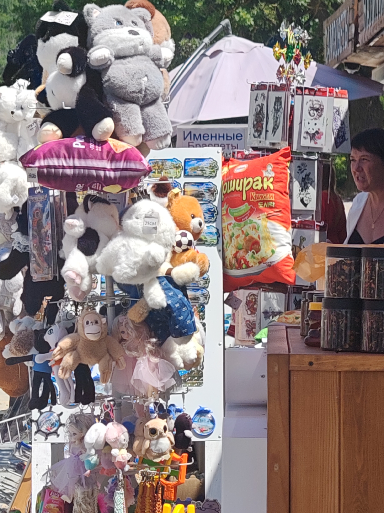 The image captures a close-up of a market stall displaying an assortment of plush toys and souvenirs, likely in the city of Sukko, Russia, given the context of the other provided images. In the foreground, a variety of stuffed animals are visible, including teddy bears, a panda, a monkey, and an owl, some adorned with clothing like dresses and overalls. Behind the toys, a large, red bag of instant noodles with Cyrillic text is prominently displayed. To the right, a woman with short dark hair is partially visible, looking to her right with a slight smile. She is wearing a white cardigan over a dark top. The stall also features shelves with small packaged items, possibly temporary tattoos, and jars filled with dried herbs or spices. The background shows a white umbrella, greenery, and parts of wooden structures, suggesting an outdoor market setting.