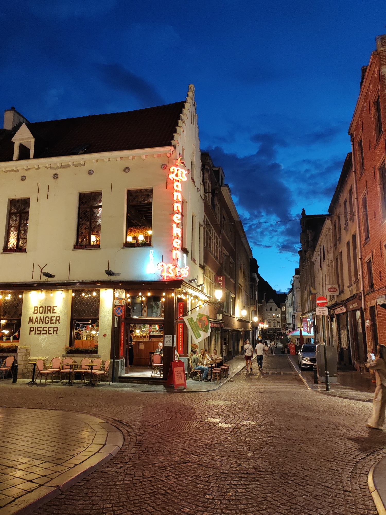 The image depicts a street scene in Brussels, Belgium, at dusk. The narrow street is paved with cobblestones, and lined with traditional European buildings. On the left, a building with cream-colored facade features a neon sign that reads "Manger Pisser". There are tables and chairs set up outside the establishment. The building on the right is made of red brick and also has illuminated windows and signage. Several people are visible walking along the street, some dining at outdoor tables. The sky is a deep blue with some clouds. The overall atmosphere suggests a lively evening in a historic European city.