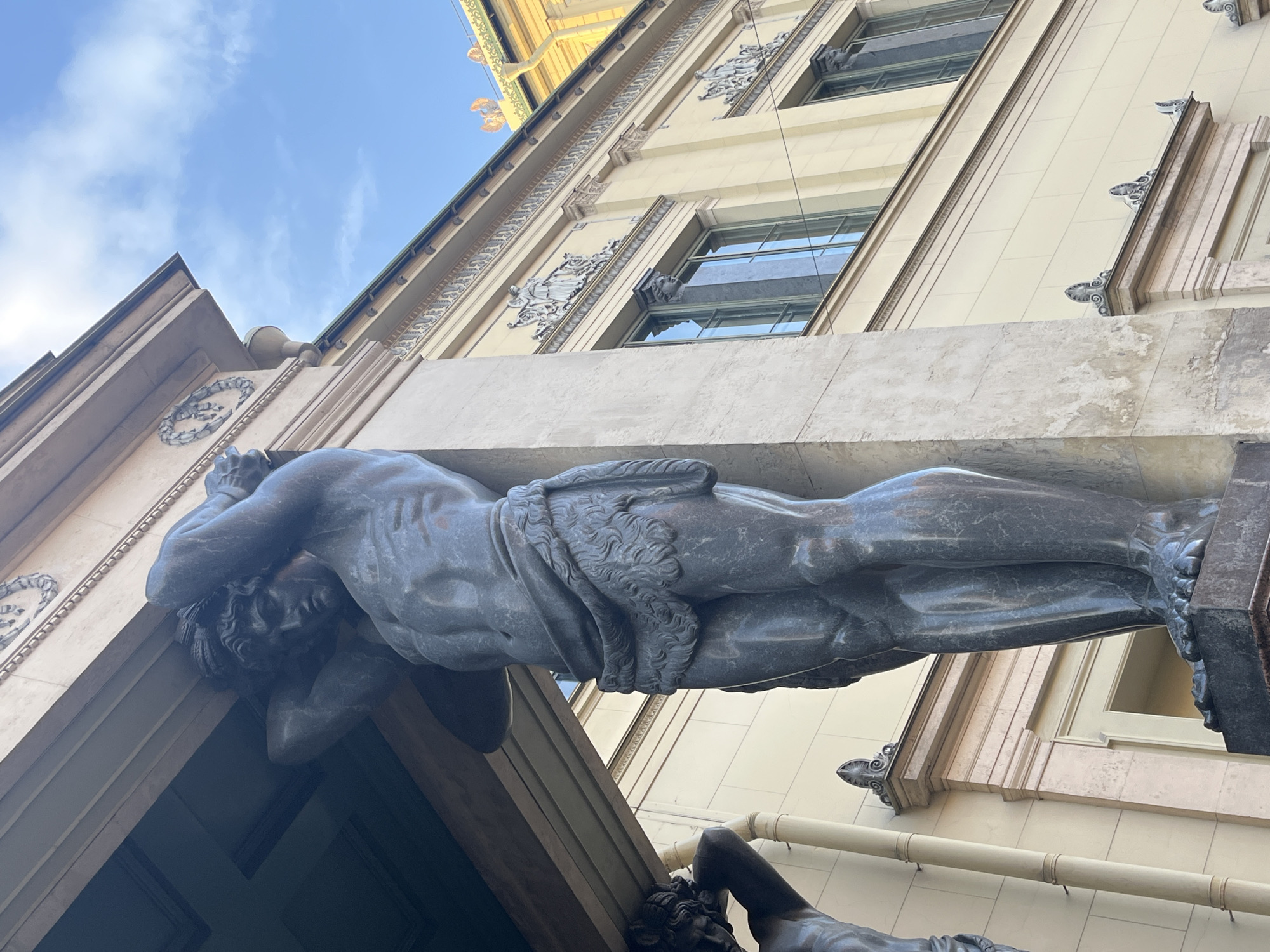 The image shows a close-up, low-angle view of one of the Atlas sculptures adorning the portico of the New Hermitage Museum in Saint Petersburg, Russia. The statue depicts a muscular, nude male figure in a strained posture, seemingly supporting a massive stone structure above. The sculpture is made of dark gray stone. Architectural details of the museum's facade are visible in the background, including windows, decorative moldings, and sections of the building's light-colored exterior. A blue sky can be seen in the upper left corner of the frame. The viewer is looking upward towards the Atlas figure.